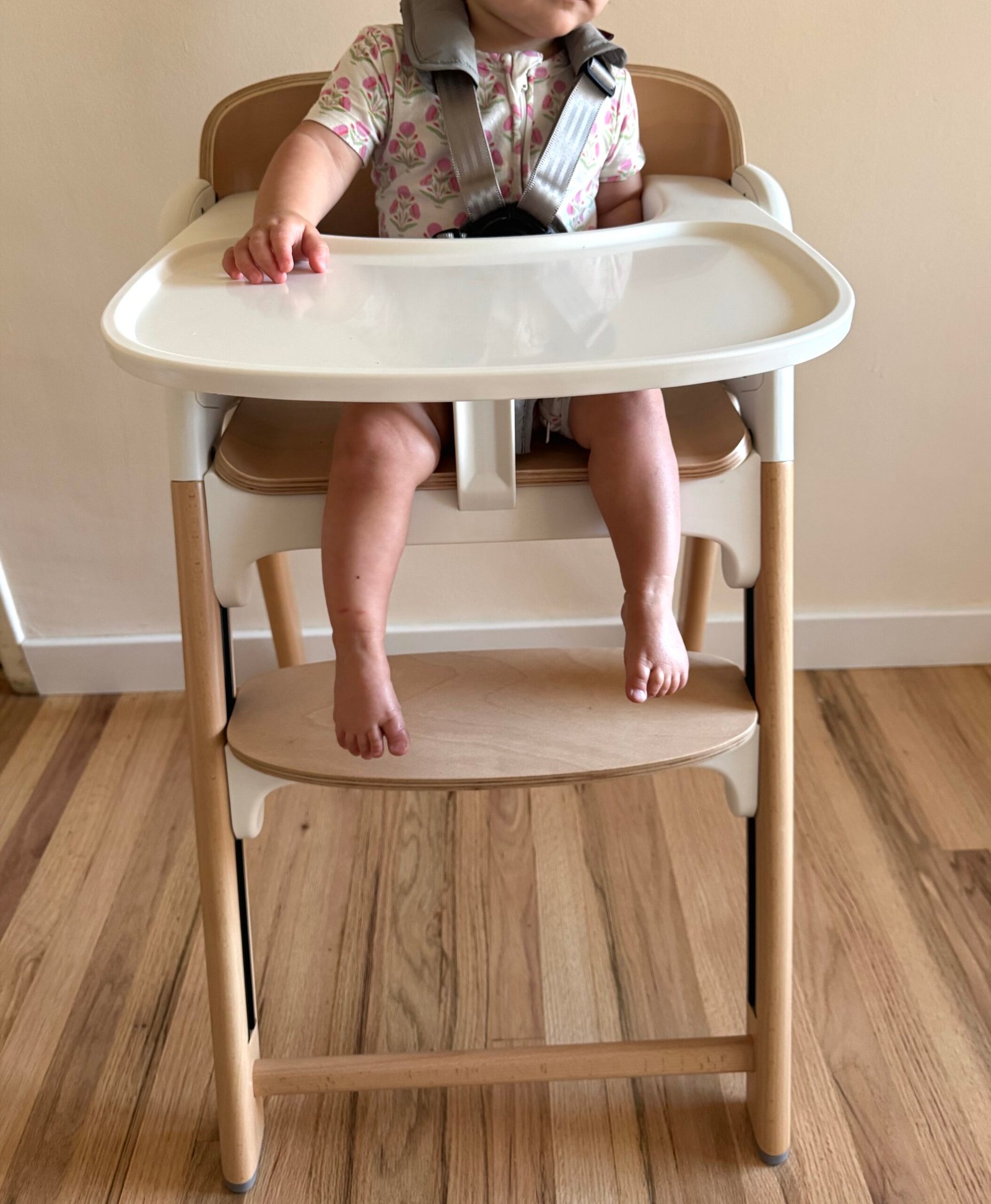 A young child sits in a wooden high chair with a white tray, wearing a floral-patterned shirt and a safety harness, feet resting on the footrest.