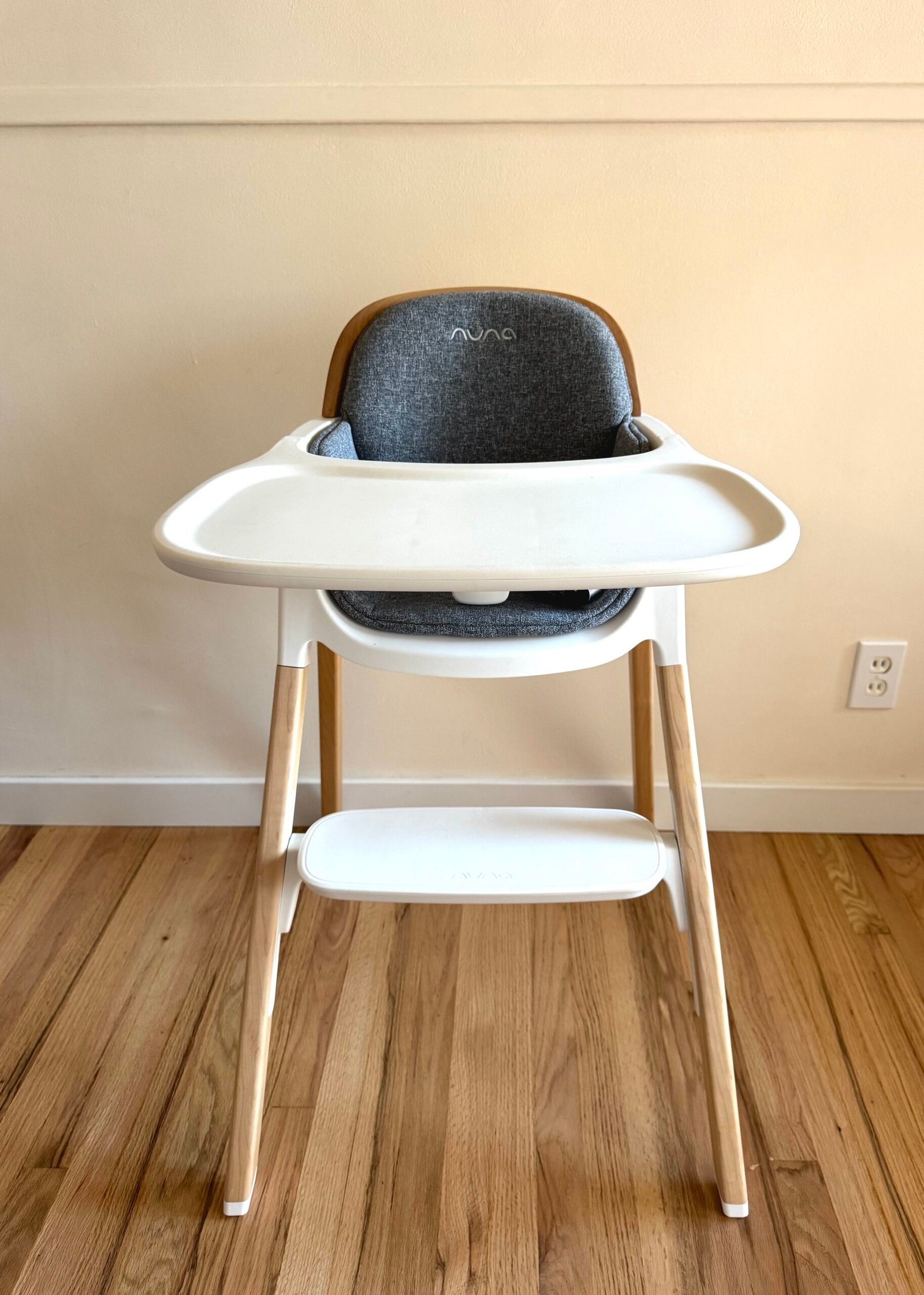 A modern high chair with a white tray and wooden legs stands on a hardwood floor against a beige wall with an electrical outlet visible on the right.