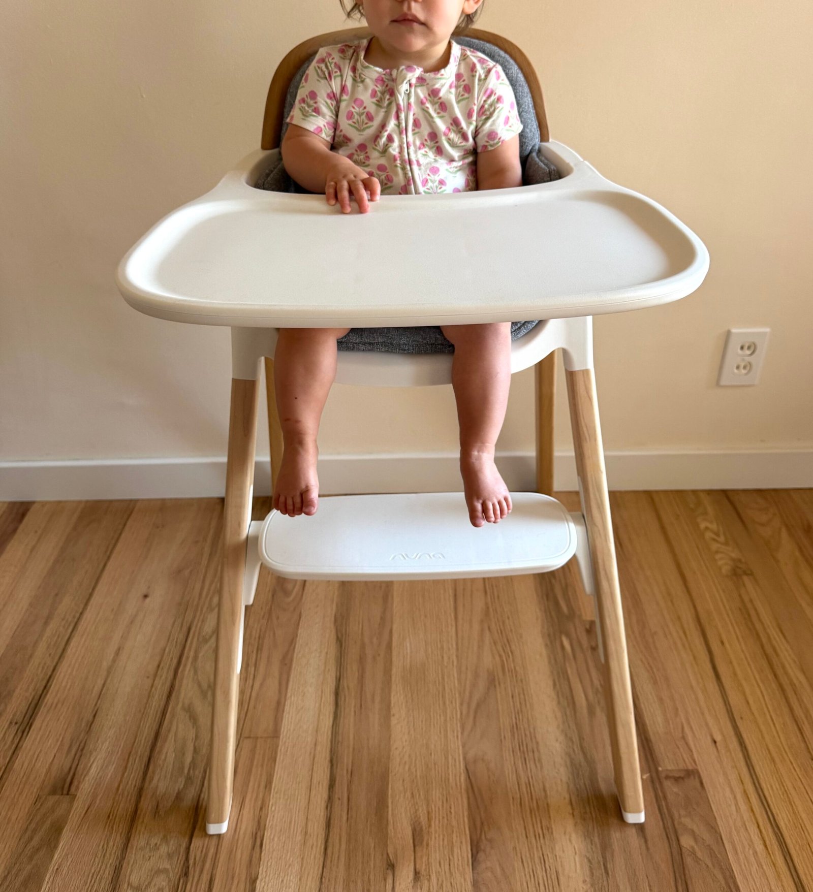 A young child sits in a high chair with a white tray and wooden legs on a wooden floor, facing the camera.