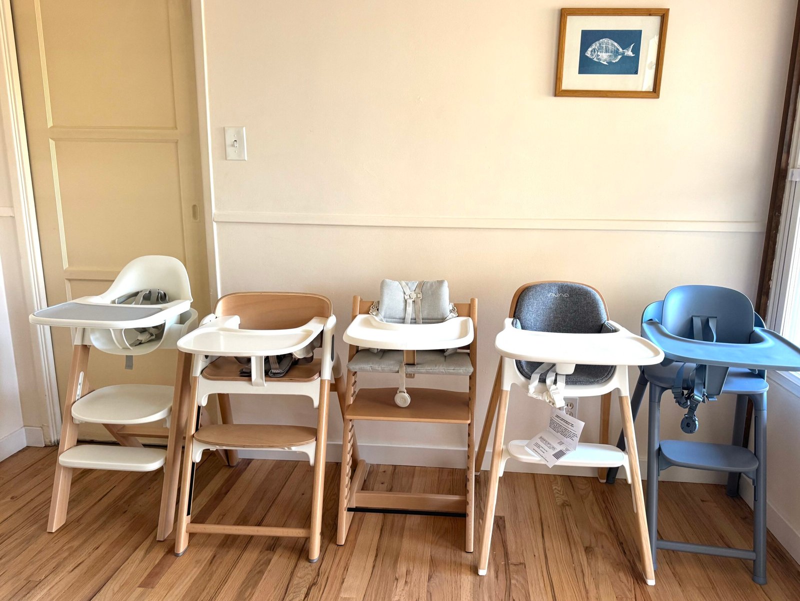 Four modern high chairs with wooden legs and plastic seats in white, gray, and blue are lined up against a cream-colored wall in a room with wooden flooring.