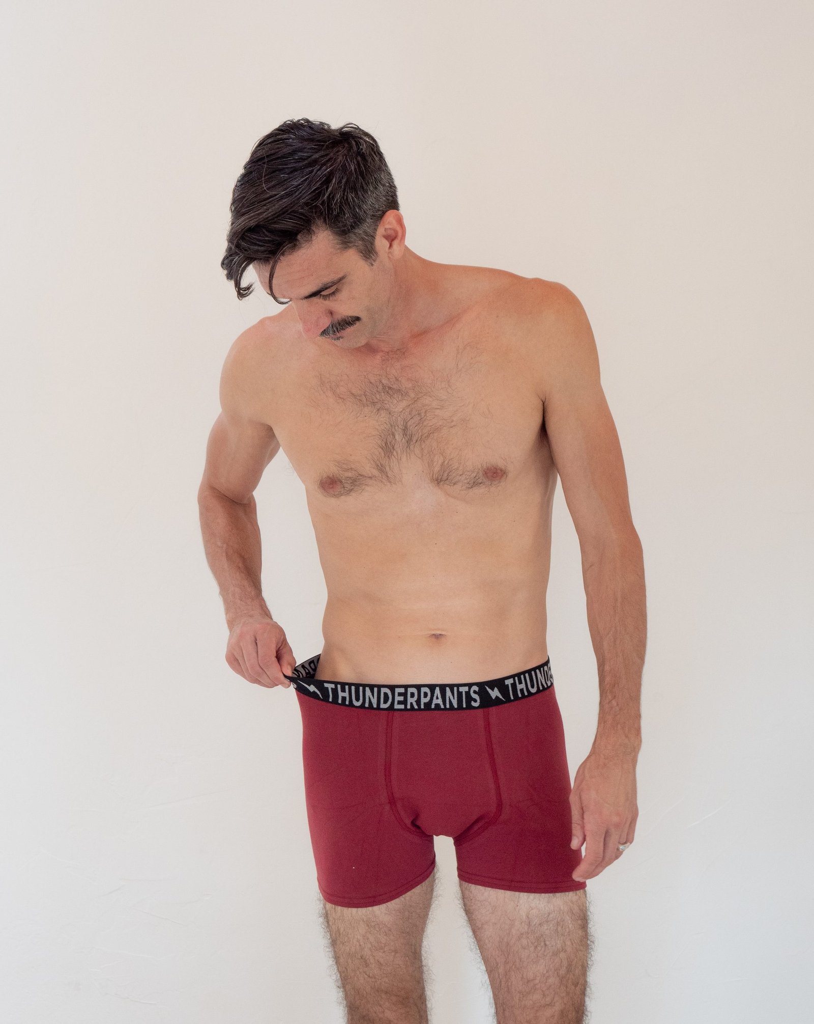 Shirtless man with a mustache looks down while adjusting the waistband of his red Thunderpants underwear against a plain background.