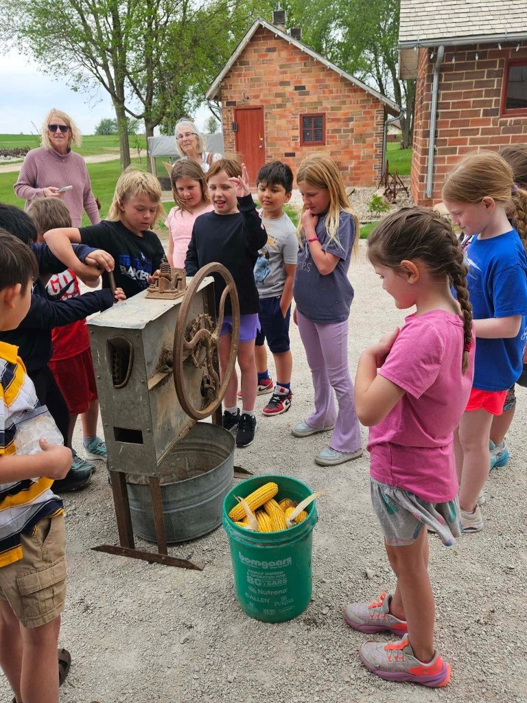 A group of children gathered around a vintage corn sheller, observing its operation while holding ears of corn. Two adults are watching from the background, with a barn and trees visible in the scenery.