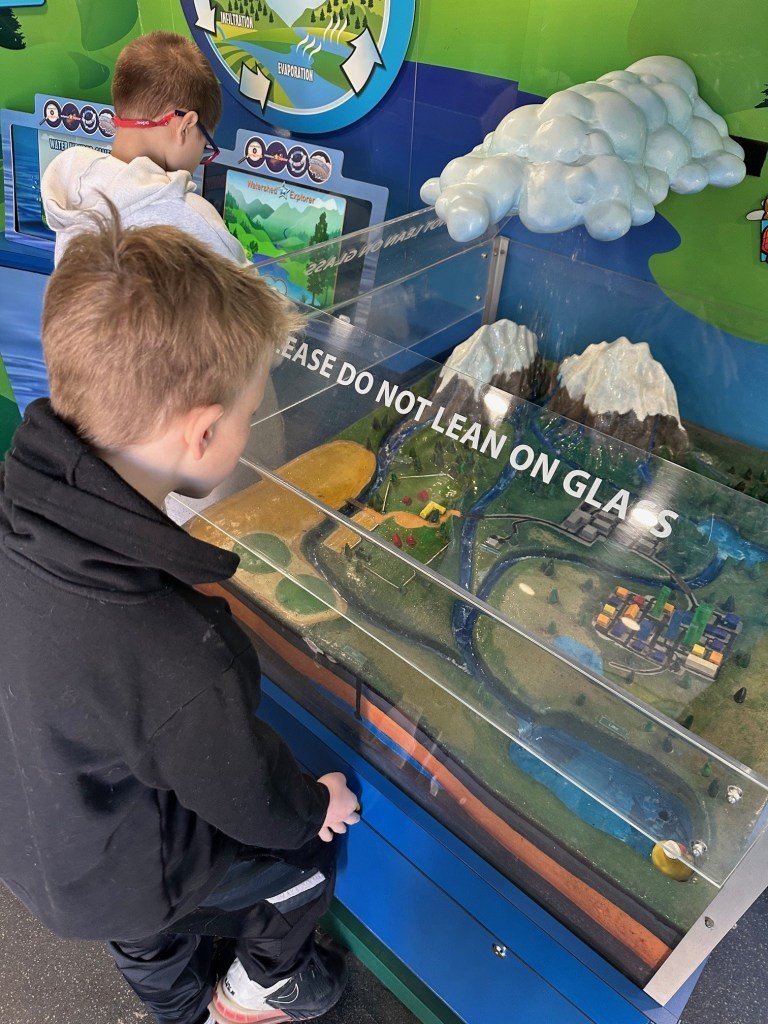Two children observe a water-themed exhibit featuring a miniature landscape with mountains, rivers, and interactive elements. A sign emphasizes not to lean on the glass.