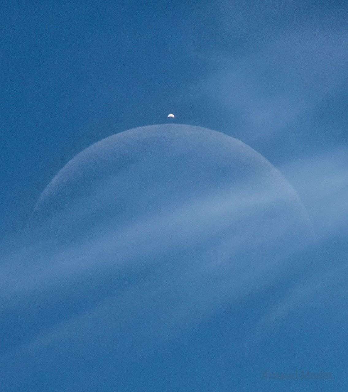 A blue sky has a light cloud crossing the middle of the image. Two crescent objects are visible through the cloud, a large faint crescent in the center and a brighter crescent above it.