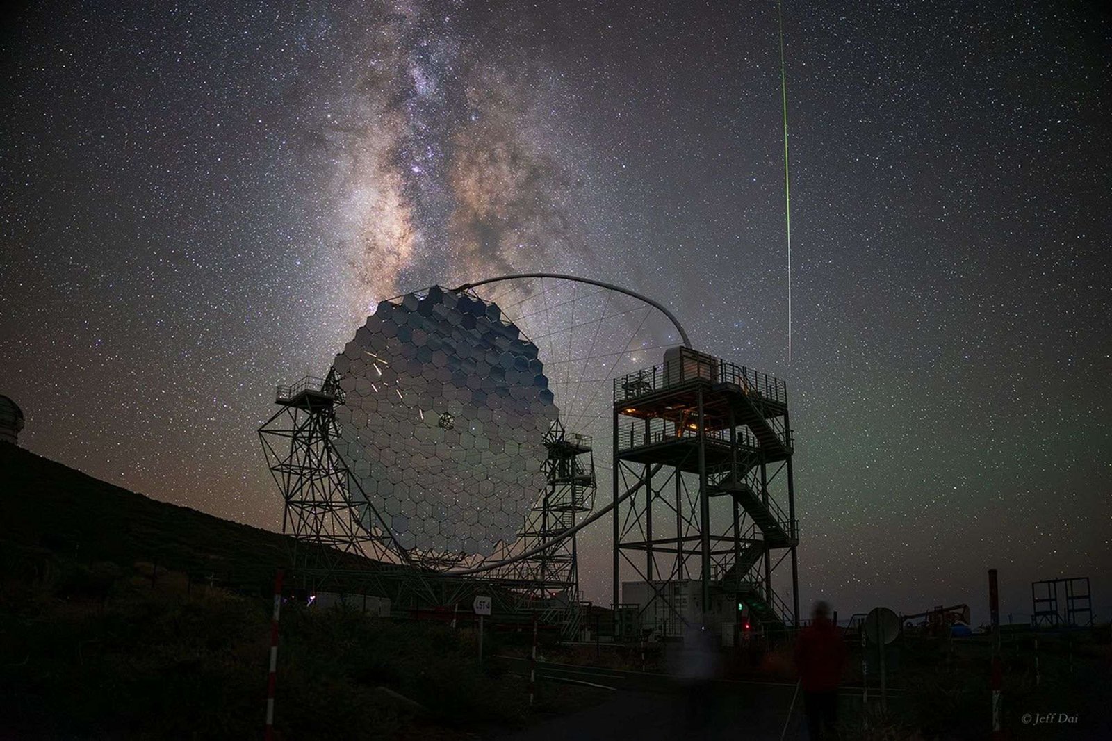 A large, segmented telescope mirror is seen against the night sky along with the Milky Way and a bright meteor.