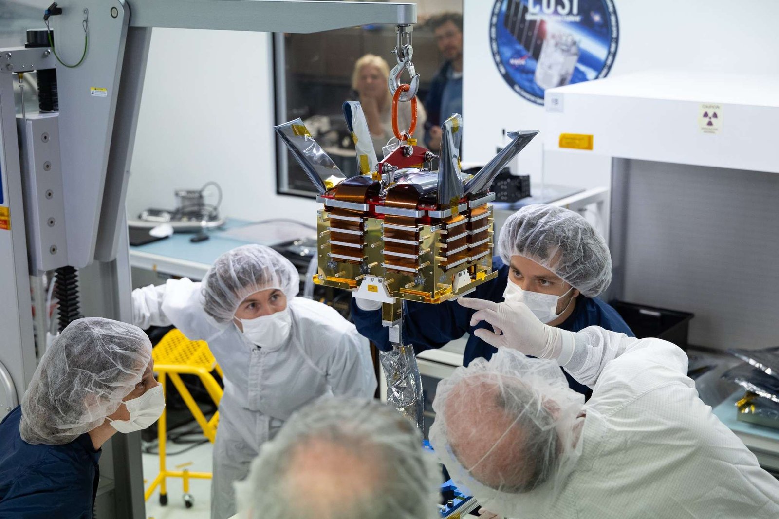 A group of five people wearing cleanroom suits, hairnets, and masks watch a small gold and silver box suspended by a small crane in a laboratory. In the background, slightly out of focus, two more people watch through a window. Next to the window is a large decal of the COSI mission patch.