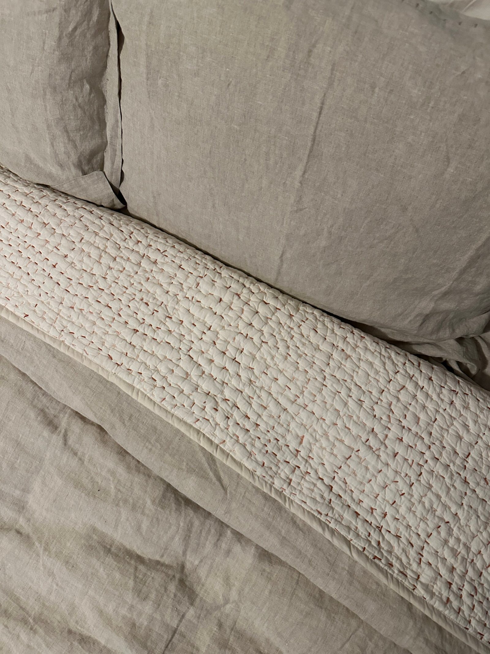 A neatly made bed with beige linen sheets, a matching pillow, and a textured white quilted blanket folded beneath the pillow.