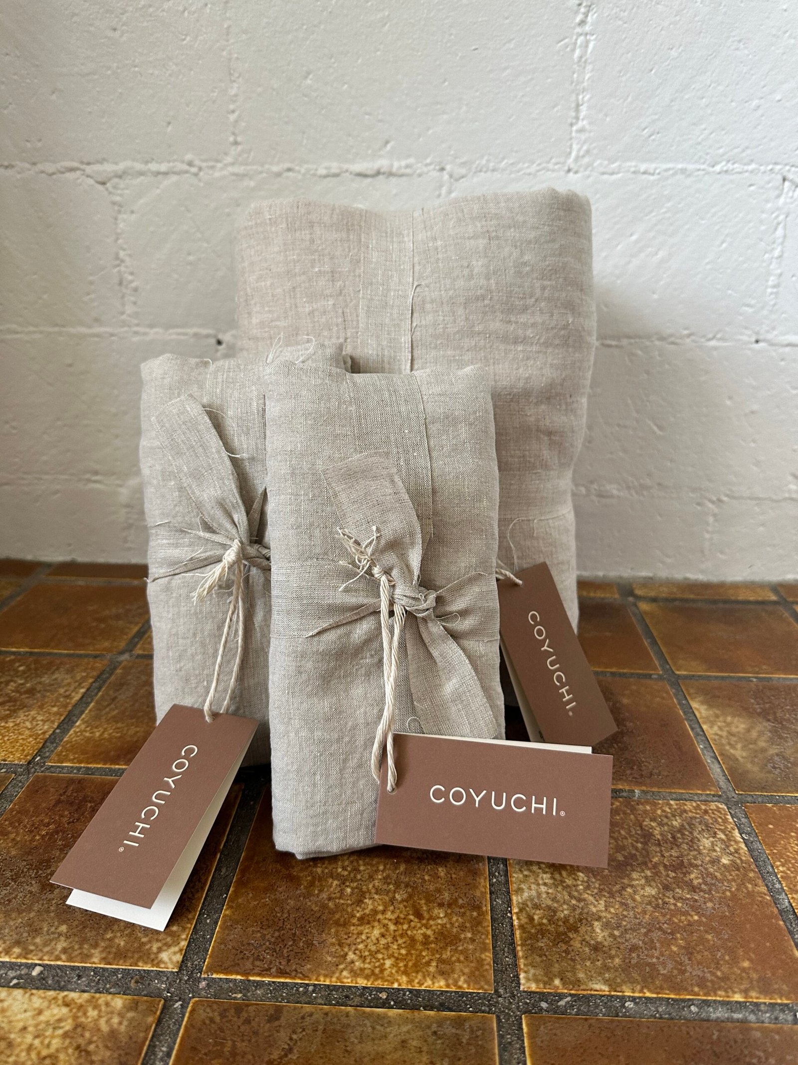 Three bundles of beige fabric tied with twine, placed on a tiled surface. Two brown tags with "COYUCHI" written on them are attached.