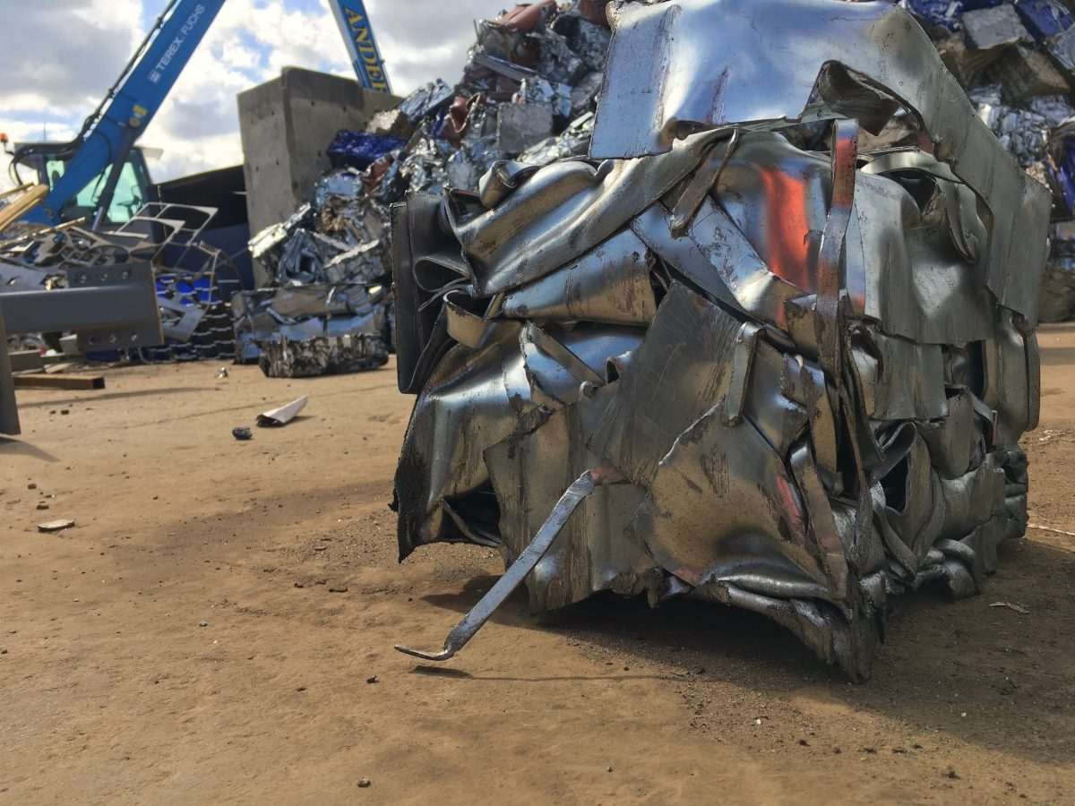 Compressed bale of scrap metal at a recycling facility, with machinery and further metal bales behind it