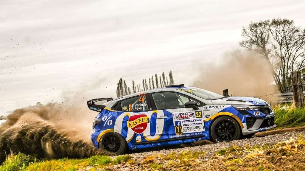 A Belgian Rally car takes a turn at high speed, with dust and dirt flying.