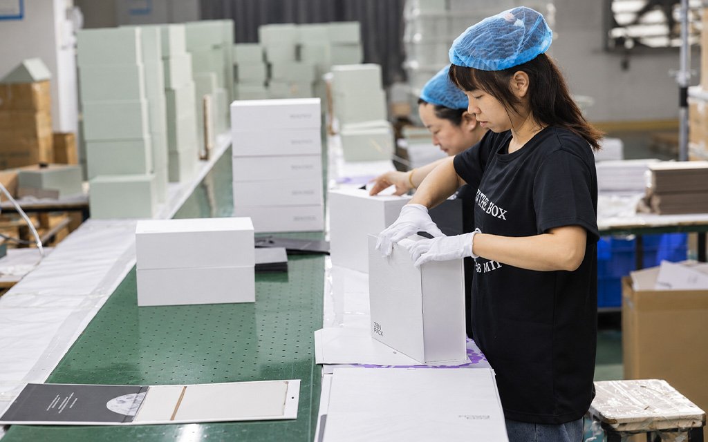 Factory workers wearing gloves and hairnets assembling white gift boxes along a production conveyor belt.