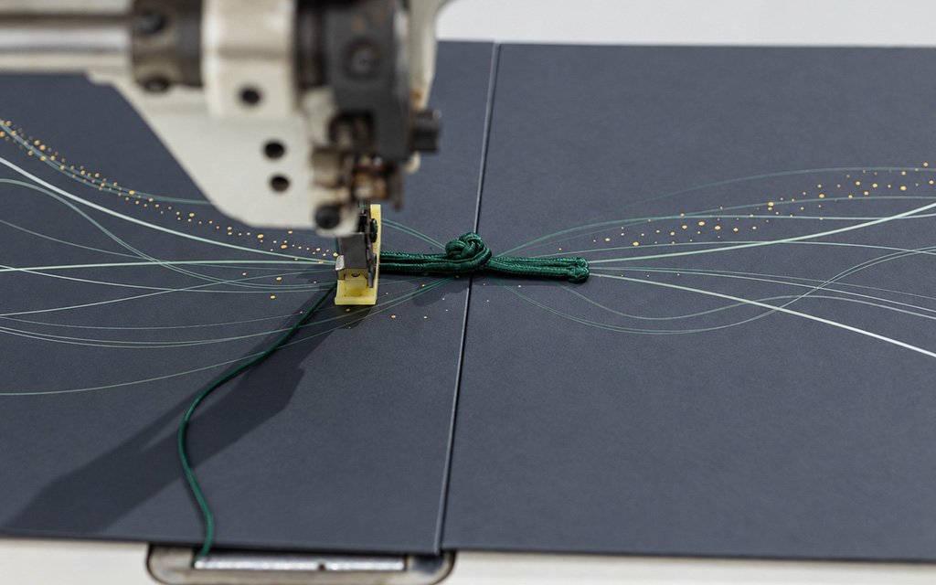 Close-up of a sewing machine attaching a green knot cord to a dark blue box panel.
