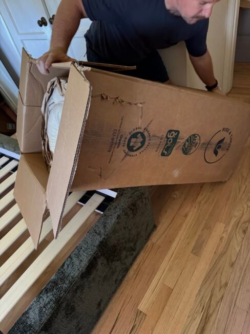 A person unboxes a rolled-up mattress onto a wooden bed frame in a bedroom with hardwood floors.