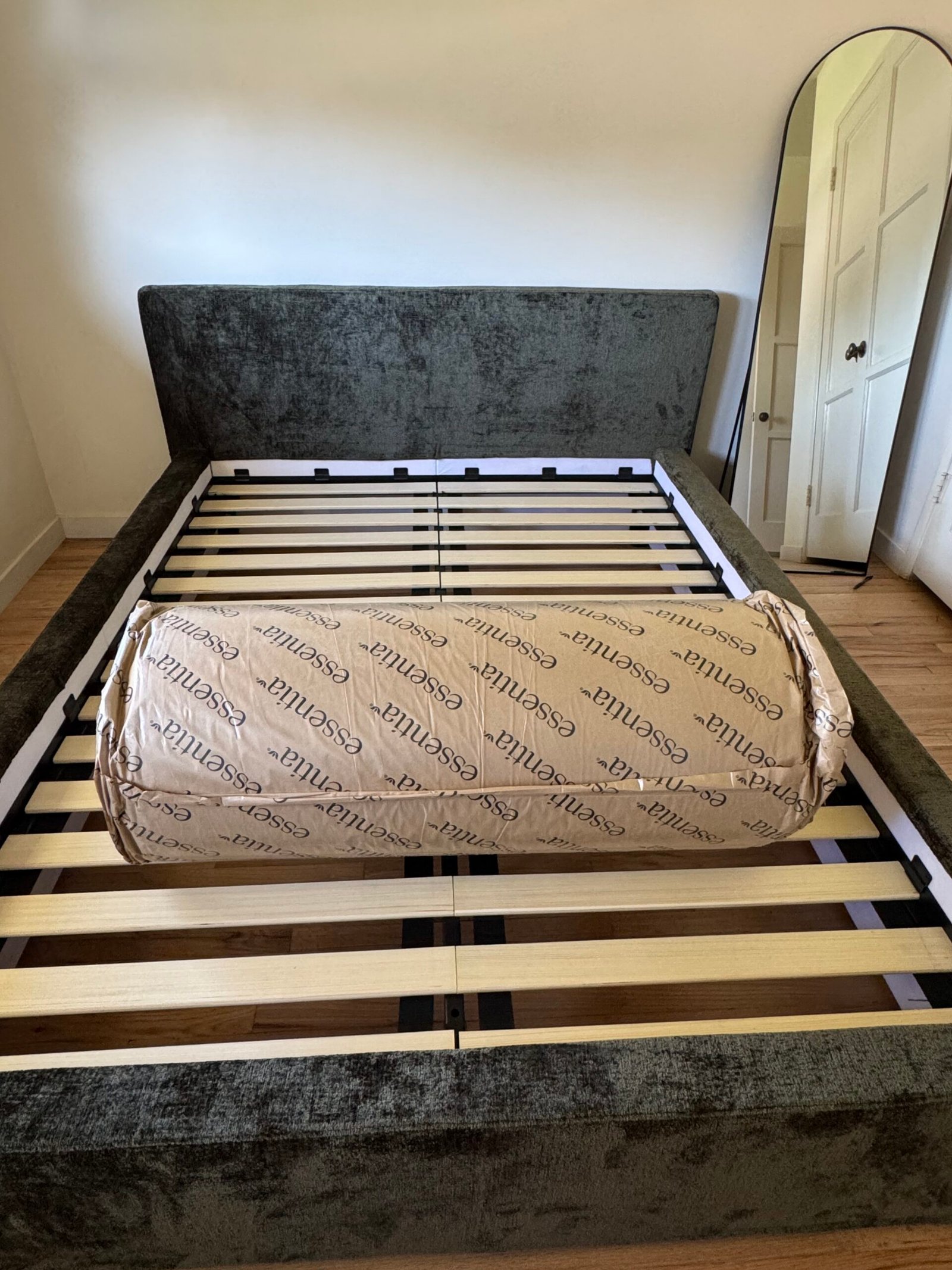 A rolled-up mattress in brown packaging labeled “essentia” sits on the slats of an empty bed frame in a bedroom with a mirror and wooden floor.