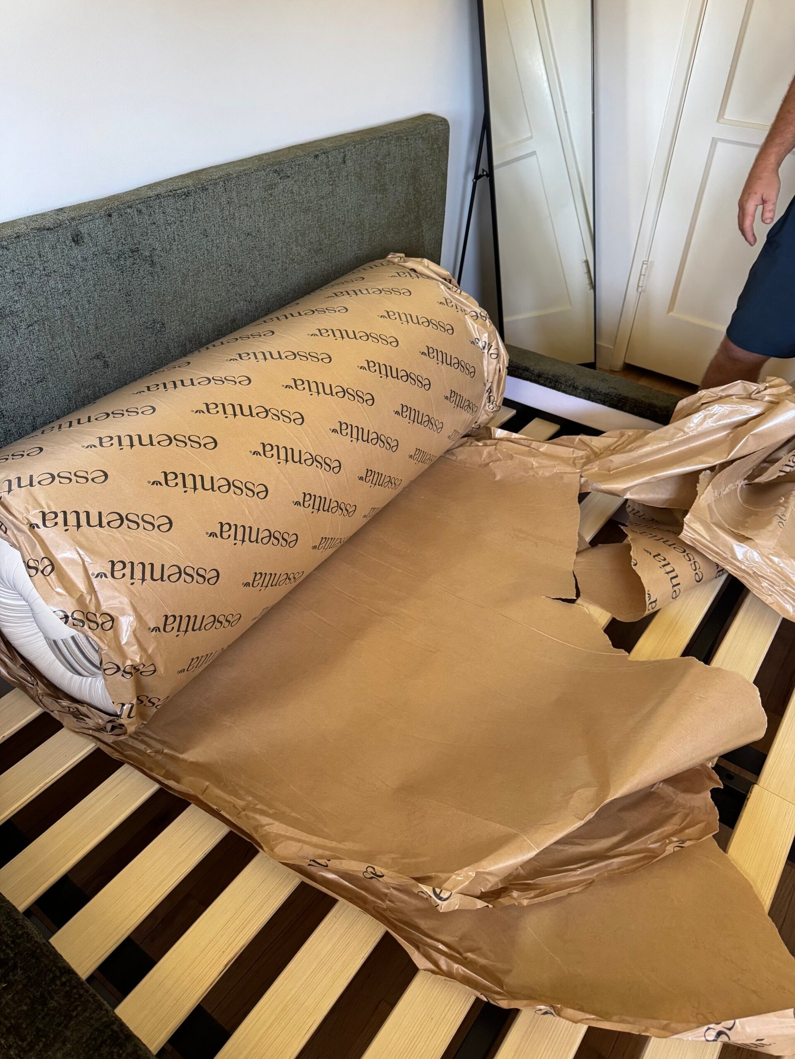 A rolled-up mattress in branded packaging is partially unwrapped and resting on a slatted bed frame, with torn brown paper nearby.