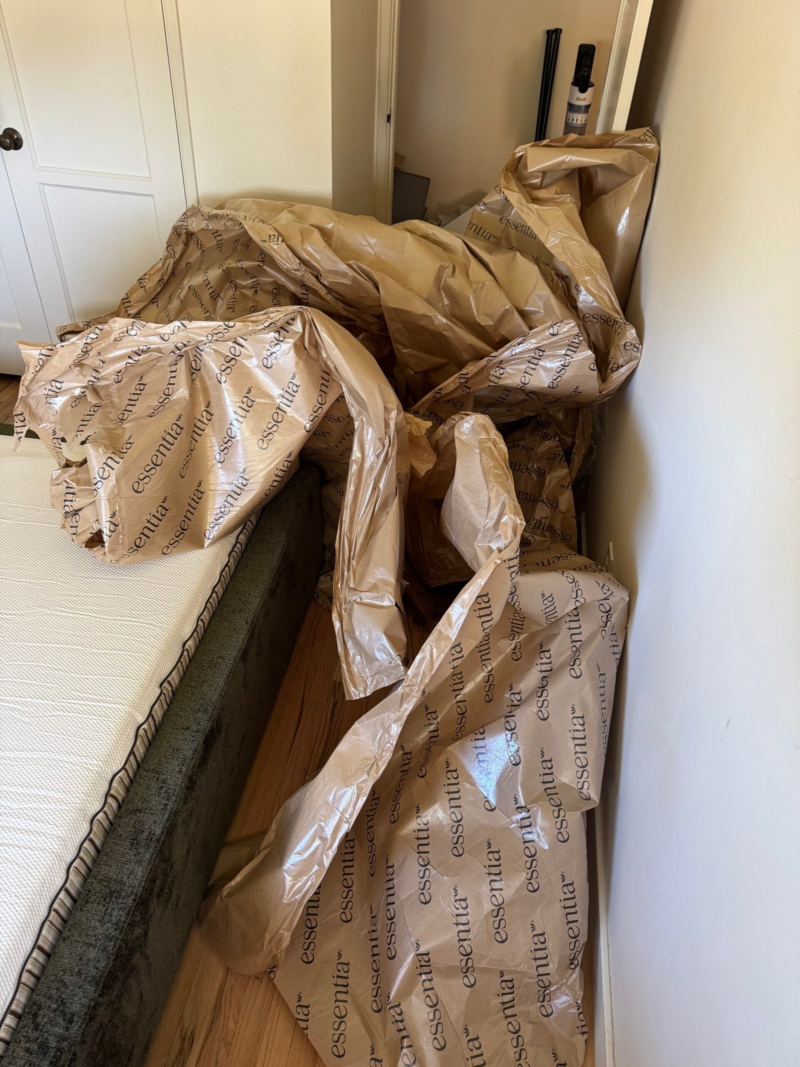 Large sheets of brown packaging paper with “essential” branding are spread across a bedroom floor next to a bed, partially covering furniture and touching a wall.