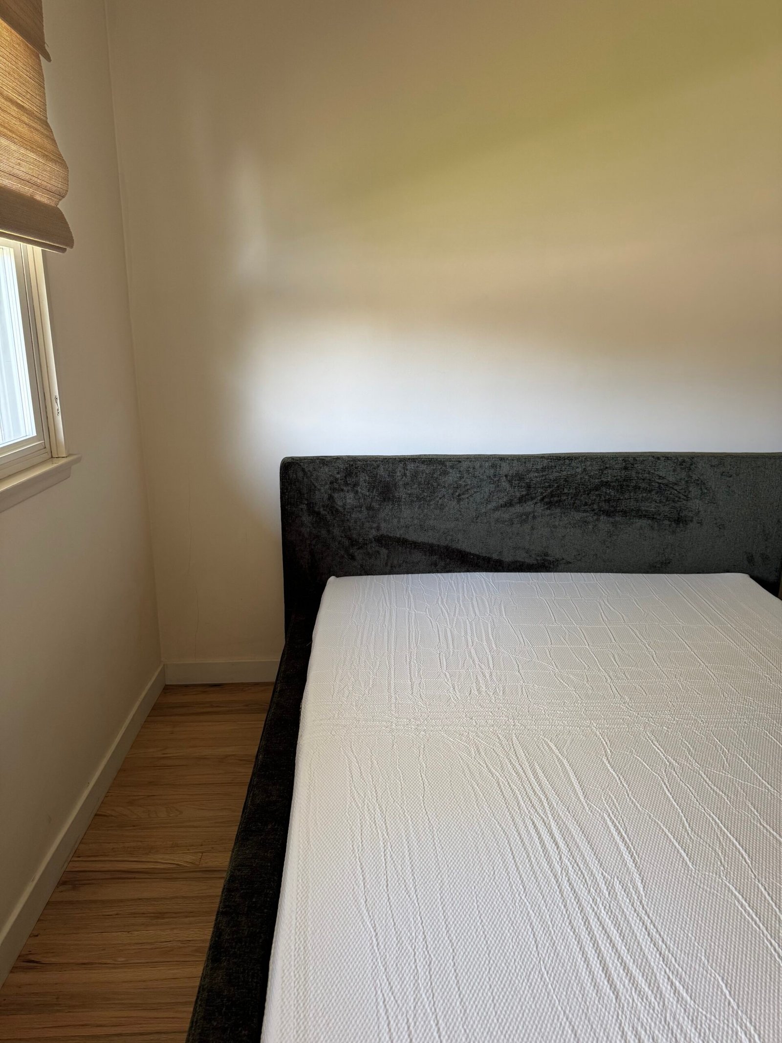 A neatly made bed with a white sheet is placed against a plain wall next to a window with blinds in a minimally furnished room with wooden flooring.