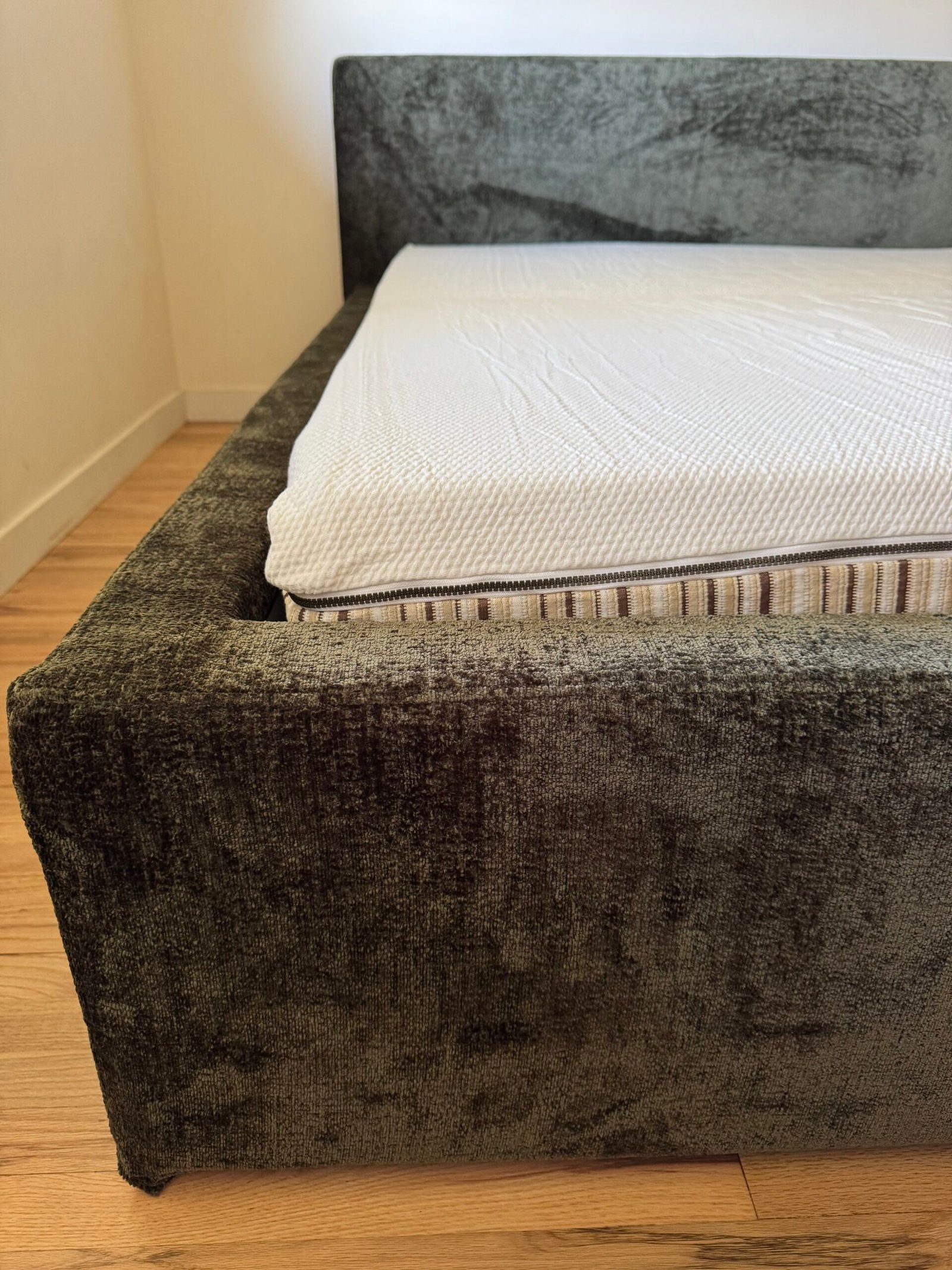 A close-up of a modern upholstered bed frame with a mattress partially covered by a textured white mattress topper, on a wooden floor.