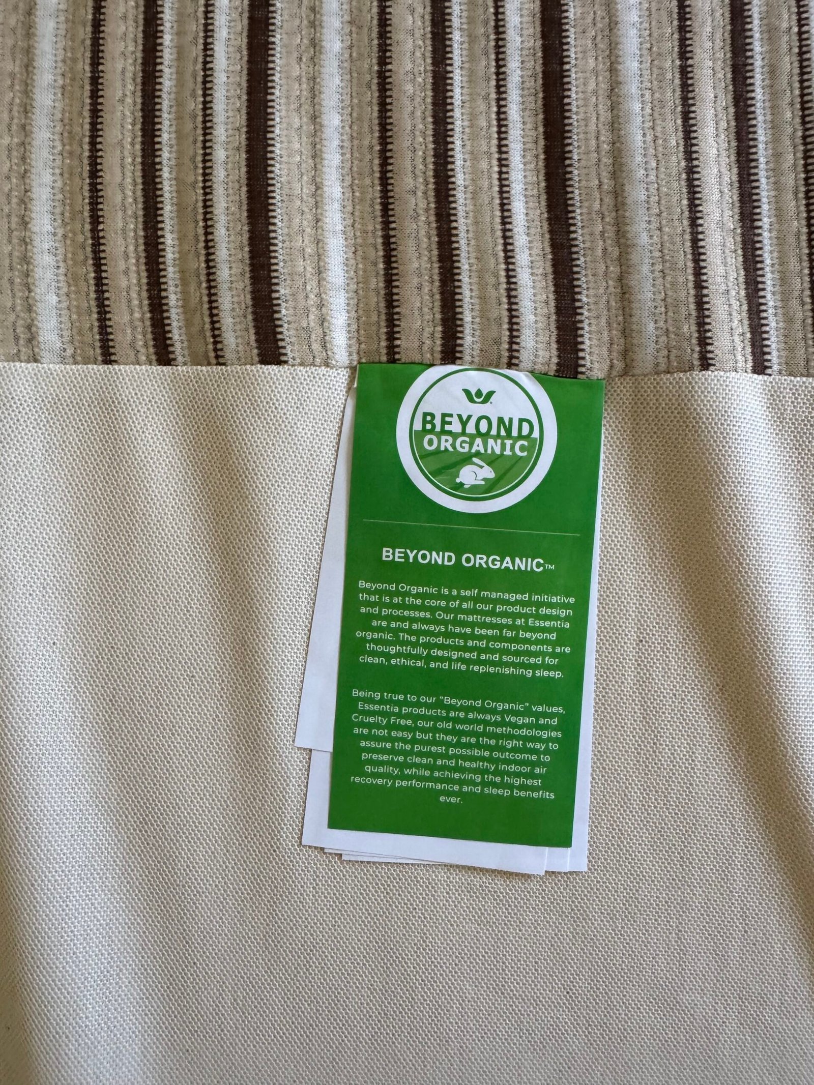 A green “Beyond Organic” label with white text is attached to beige and striped fabric, describing the initiative’s commitment to organic, ethical, and sustainable practices.