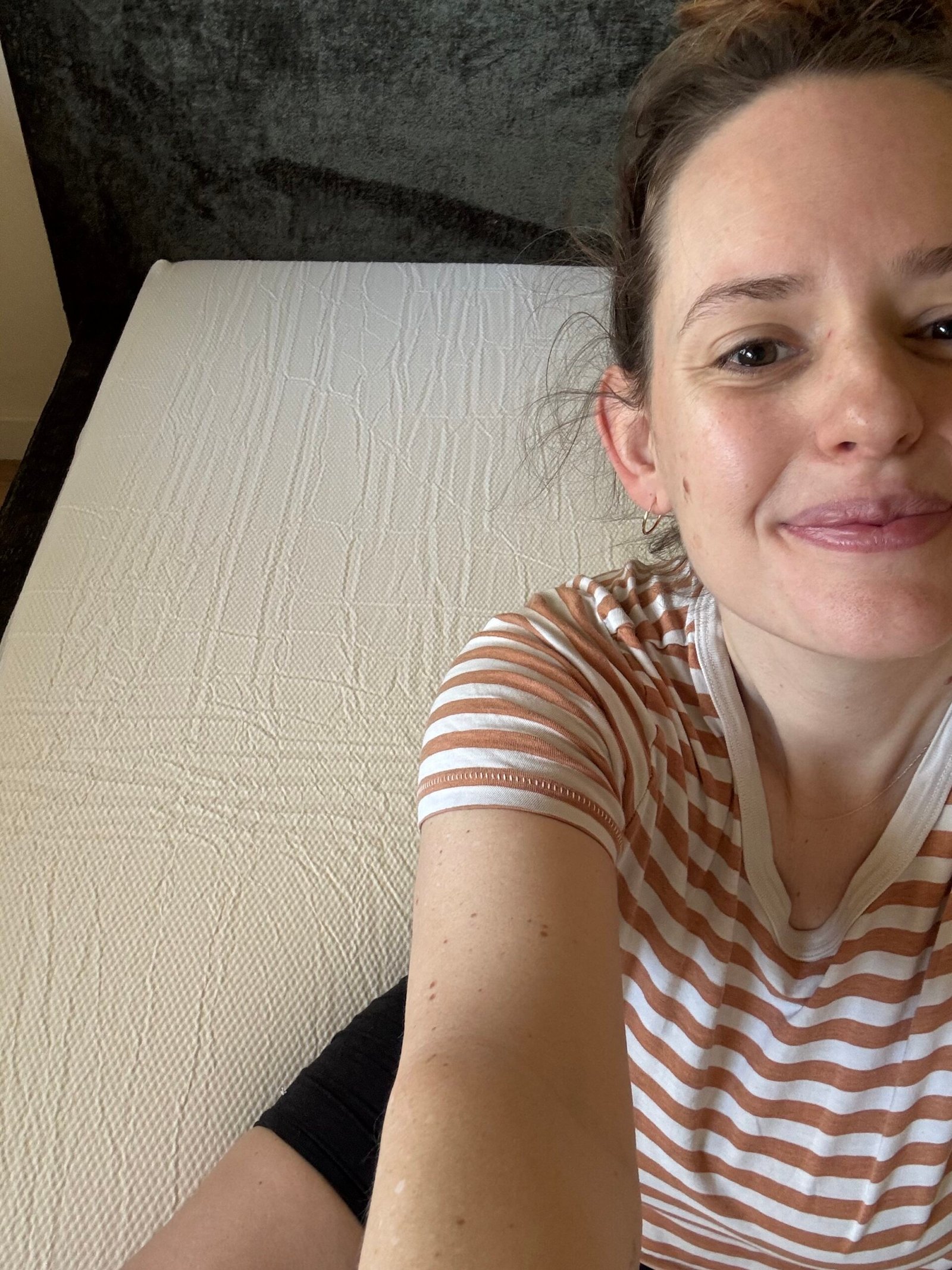 Person wearing a striped shirt sits on the edge of a bare mattress, smiling at the camera. The background includes a dark headboard.