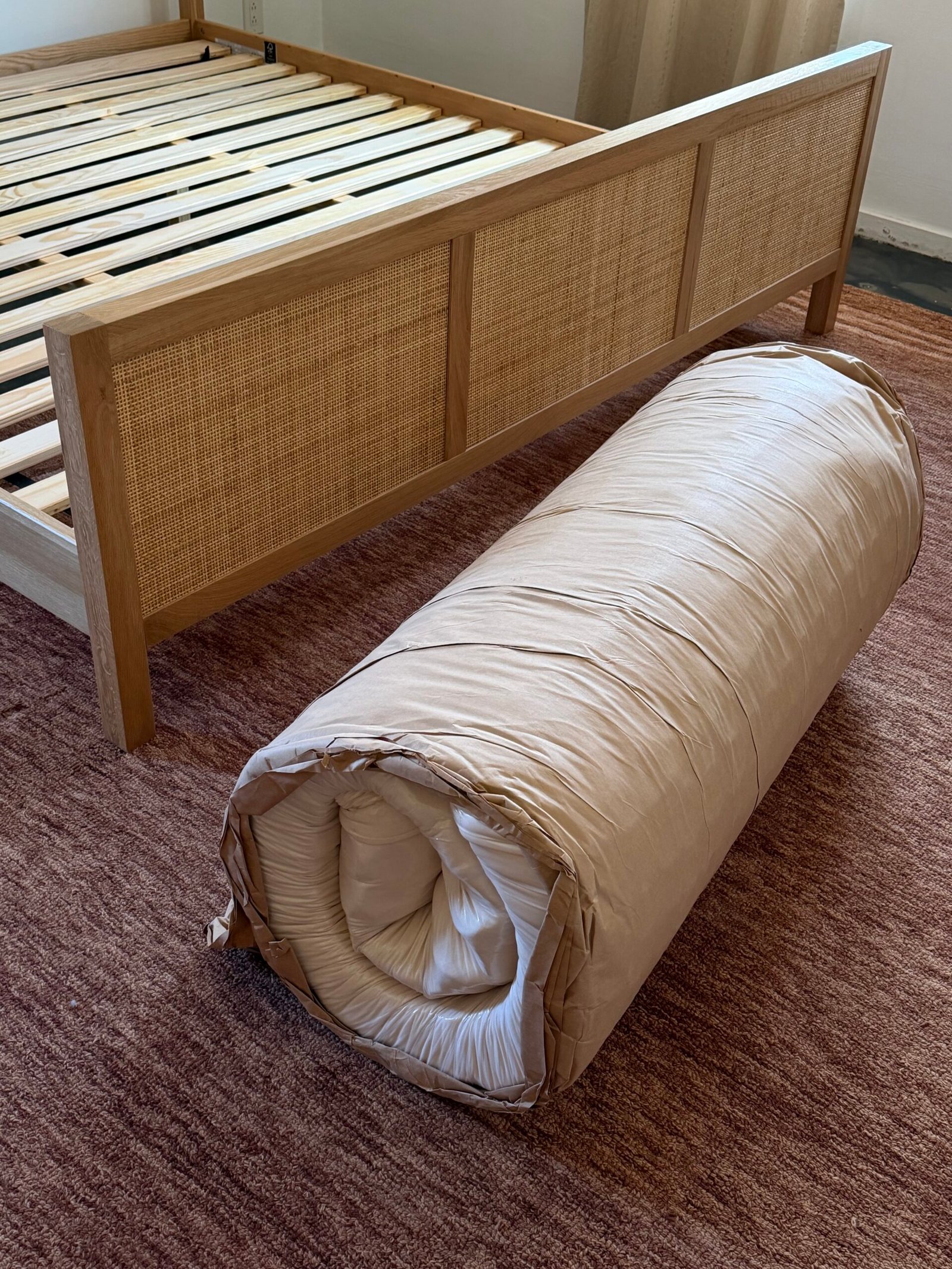 A rolled-up mattress sits on a brown rug next to a wooden bed frame with no mattress.