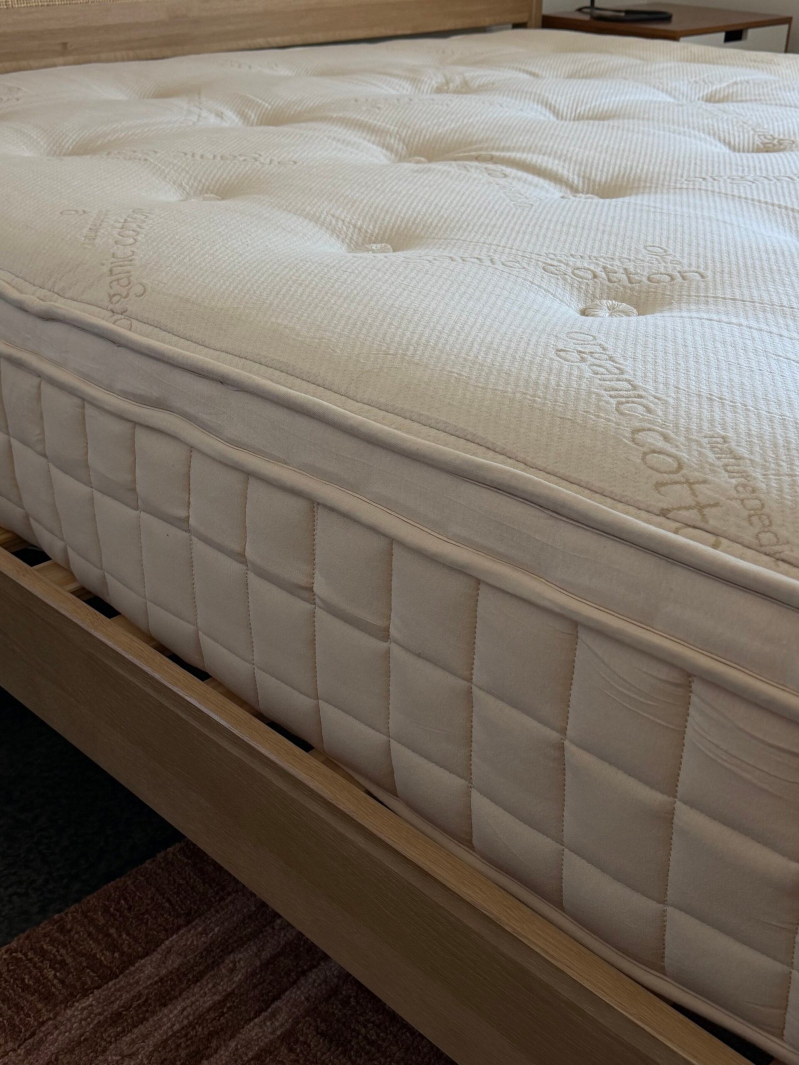 Close-up of a neatly made mattress with a quilted surface and visible stitching placed on a light wooden bed frame.