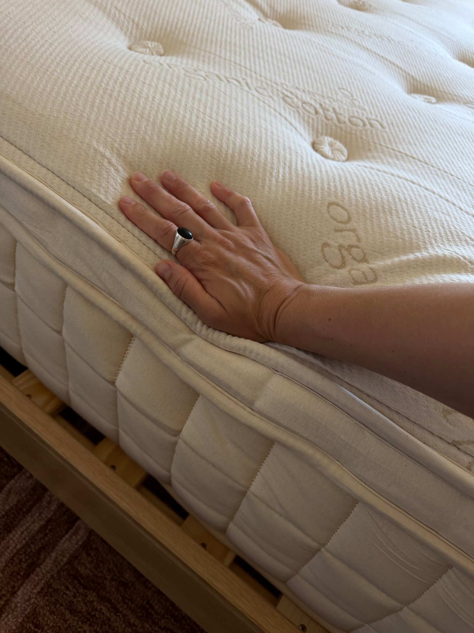 A hand pressing down on the surface of a cream-colored mattress on a wooden bed frame, showing the texture and firmness of the mattress.