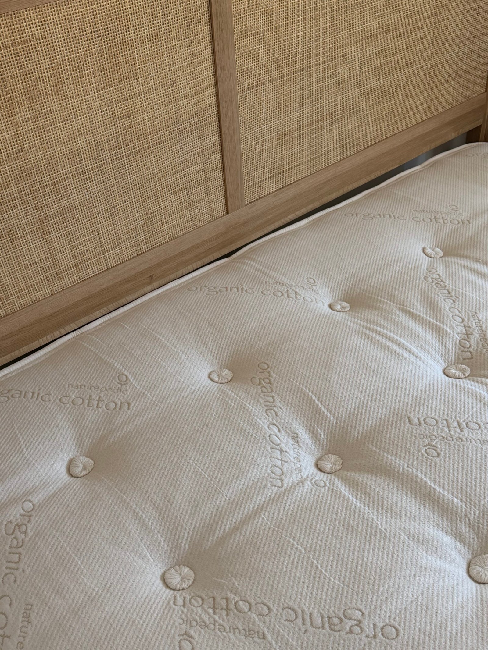 A close-up of a beige mattress labeled “organic cotton” on a wooden bed frame with a woven rattan headboard.