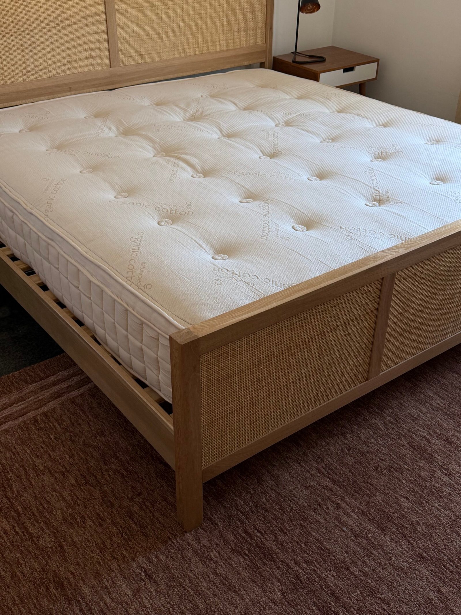A corner view of a neatly made bed with a light wood frame, rattan panels, and a white mattress on top, placed on a reddish-brown rug in a bedroom.