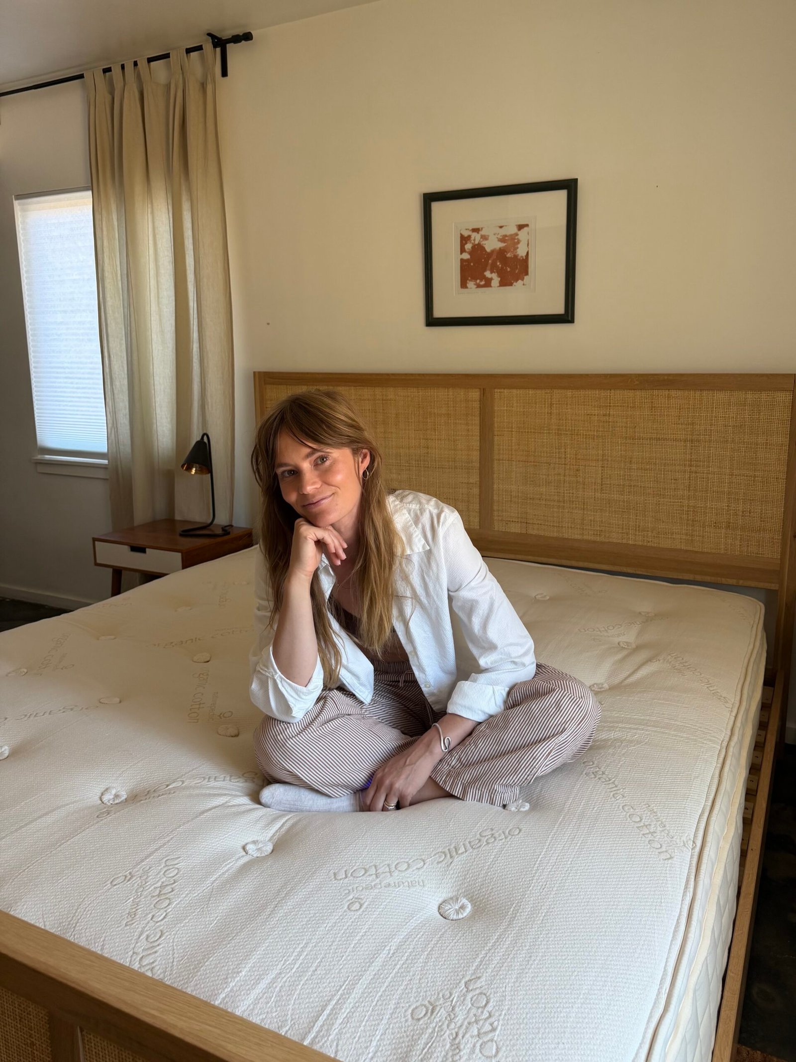 A woman sits cross-legged on an unmade bed in a simple bedroom with light walls, a framed picture, and a window with curtains.