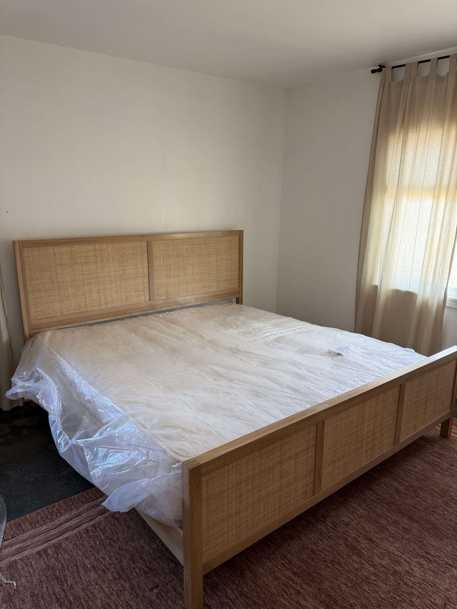 A wooden bed frame with a cane headboard and footboard stands in a room; a mattress covered in plastic sits on top. Beige curtains cover a sunlit window beside the bed.