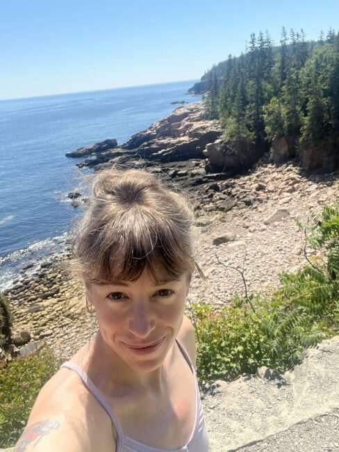 Person taking a selfie near a rocky coastline with evergreen trees, blue sky, and ocean in the background on a sunny day.