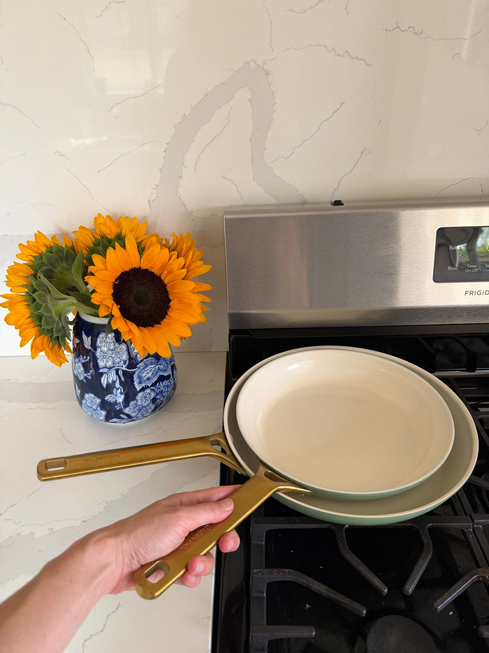 A hand holds two ceramic frying pans above a stove, with a vase of sunflowers on the counter nearby.