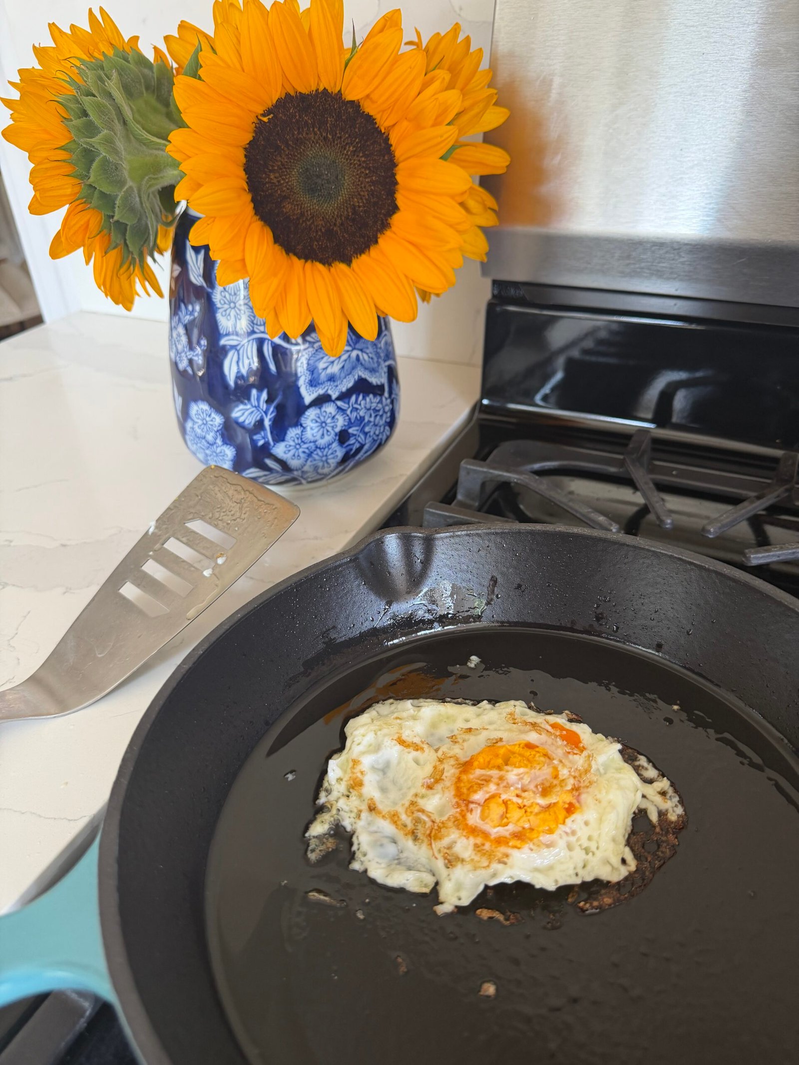 A fried egg cooking in a cast iron skillet on a stove, with a metal spatula and a blue vase of sunflowers on the white countertop nearby.