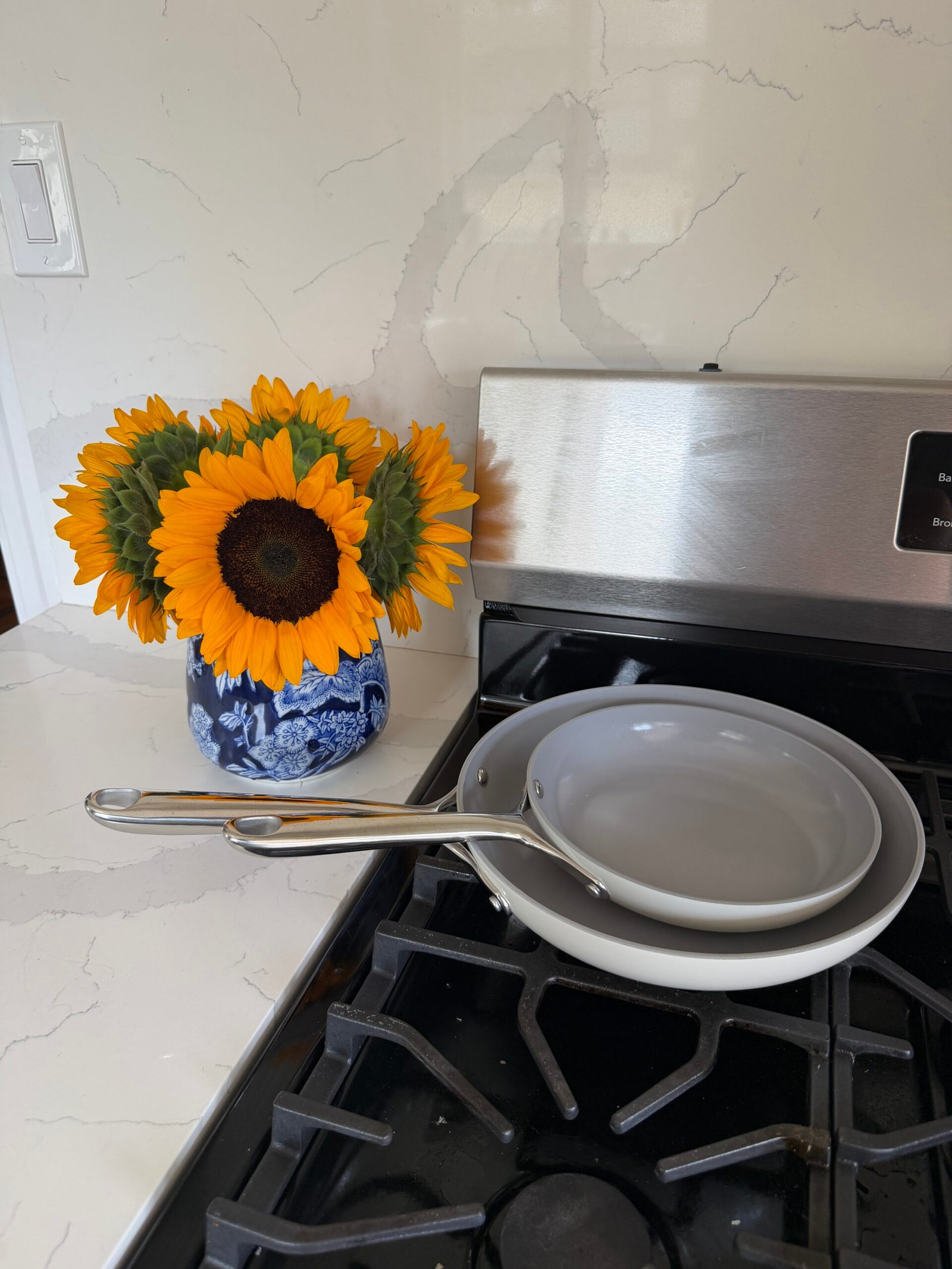 Two gray frying pans are stacked on a stove next to a blue vase with sunflowers on a white kitchen countertop.