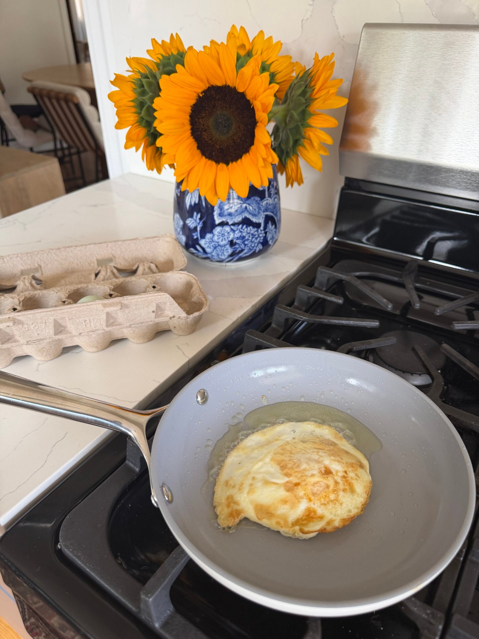 A fried egg cooks in a skillet on a gas stove, with an empty egg carton and a vase of sunflowers on the counter nearby.