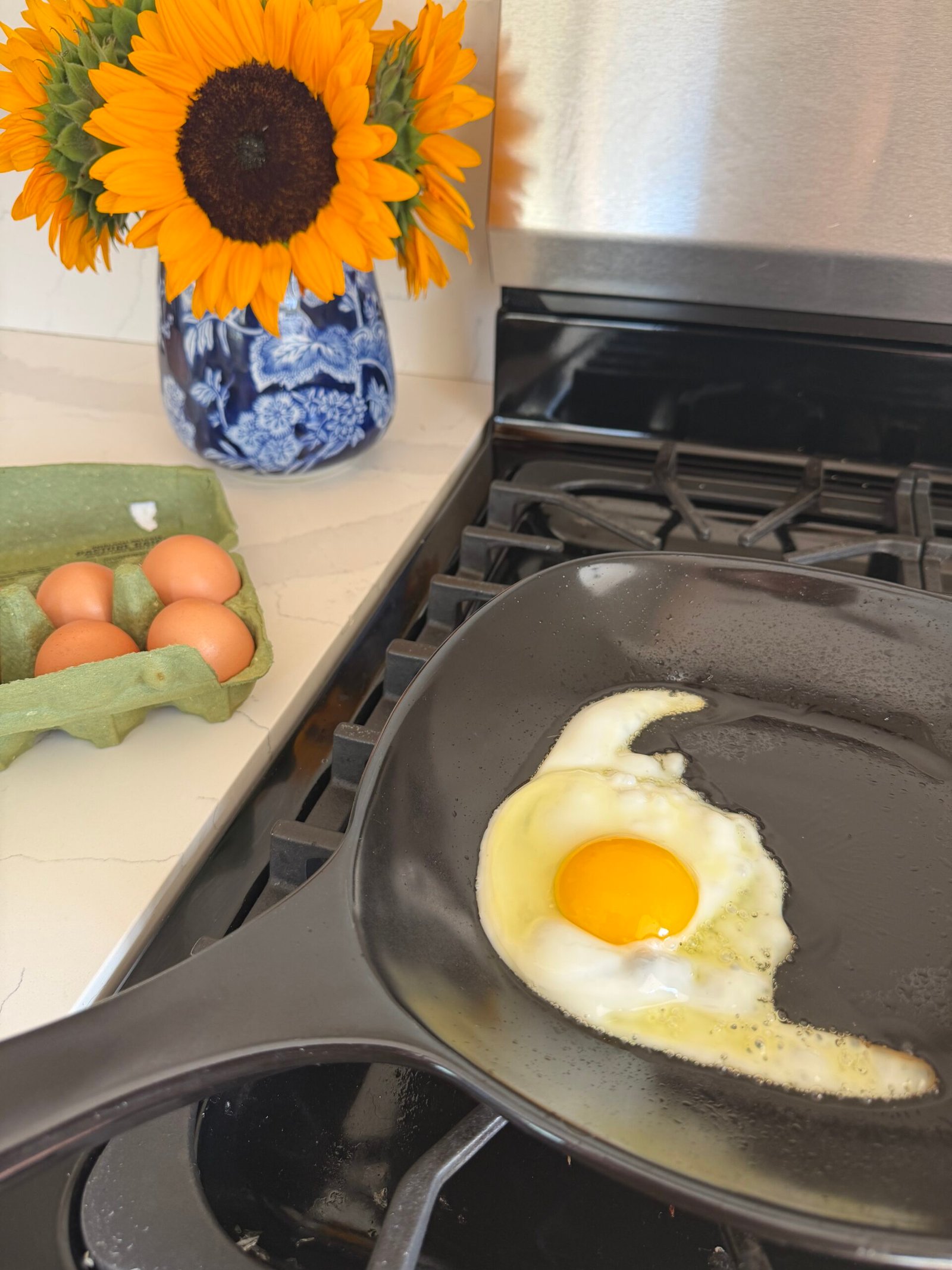 A single egg is frying in a black skillet on a stove. Next to it are three brown eggs in a carton and a vase of sunflowers on a white countertop.