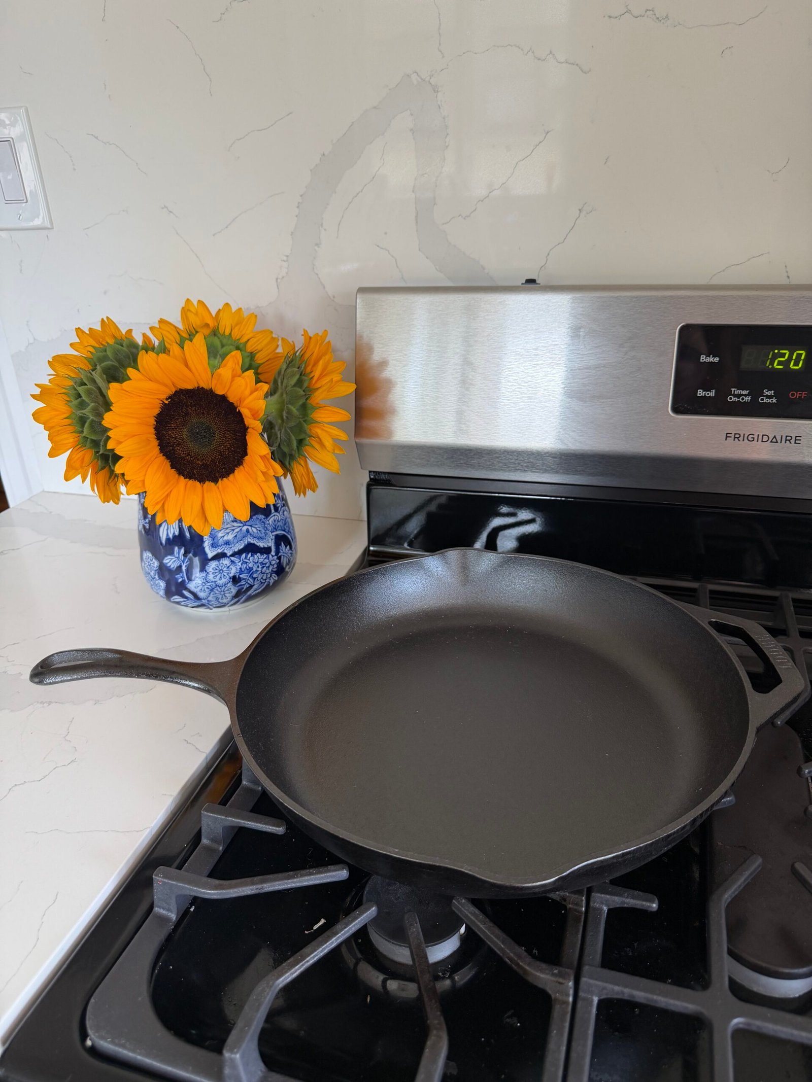A black cast iron skillet sits on a gas stove next to a blue and white vase of sunflowers.