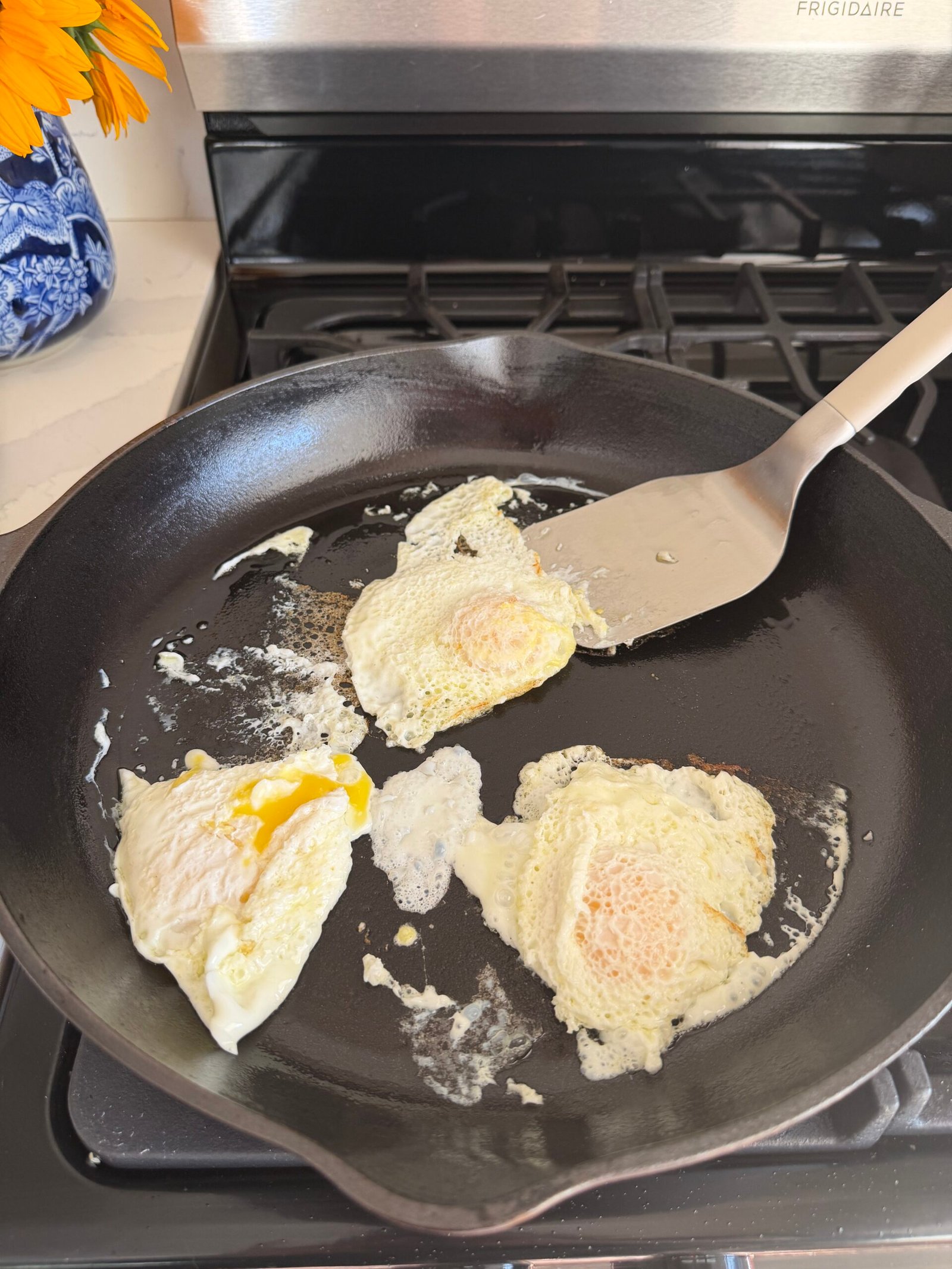 Three fried eggs are cooking in a black cast iron skillet on a stove, with a metal spatula underneath one egg.