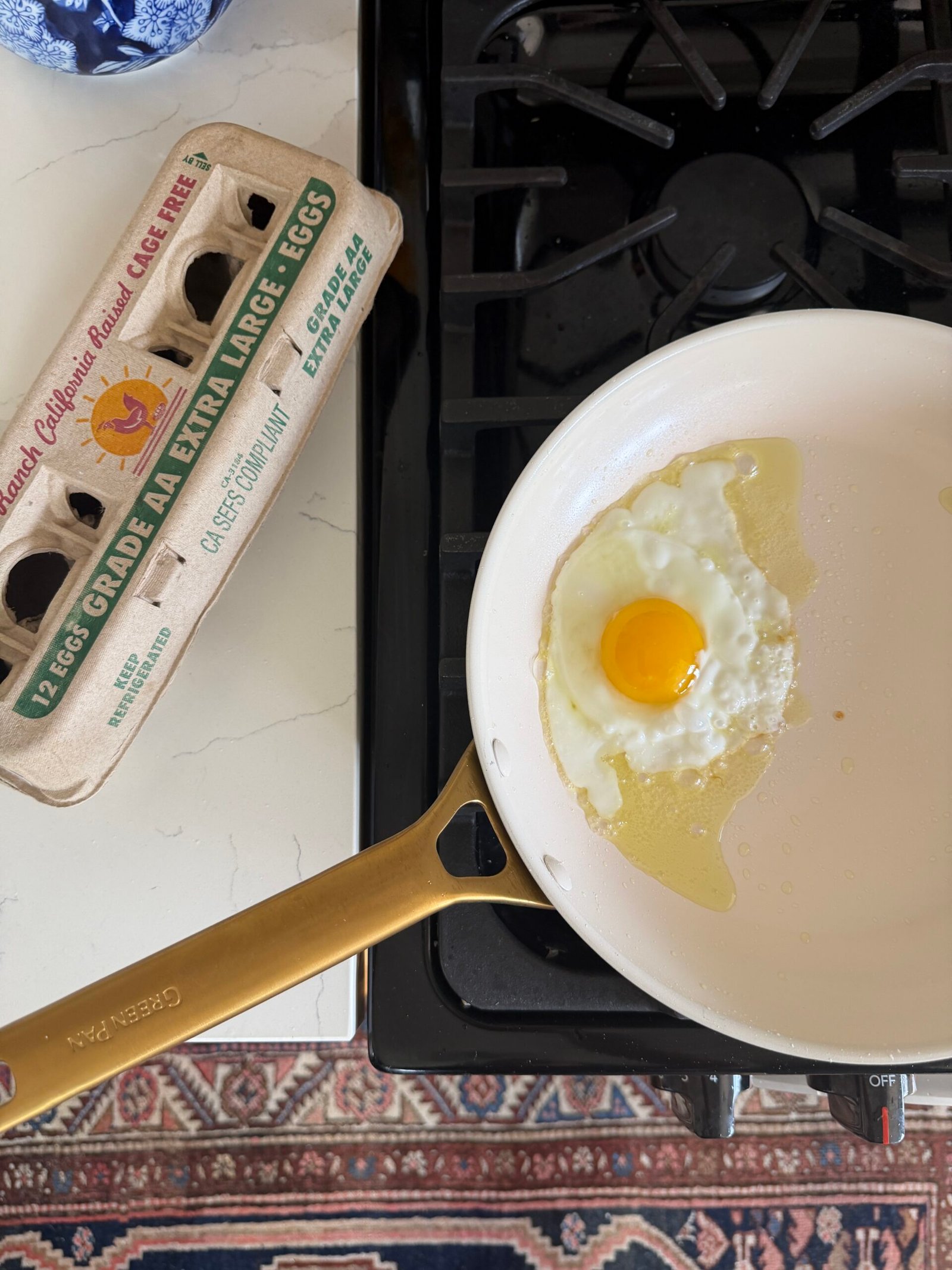 A single sunny-side-up egg cooks in a white frying pan on a stove next to an open carton of extra-large eggs.