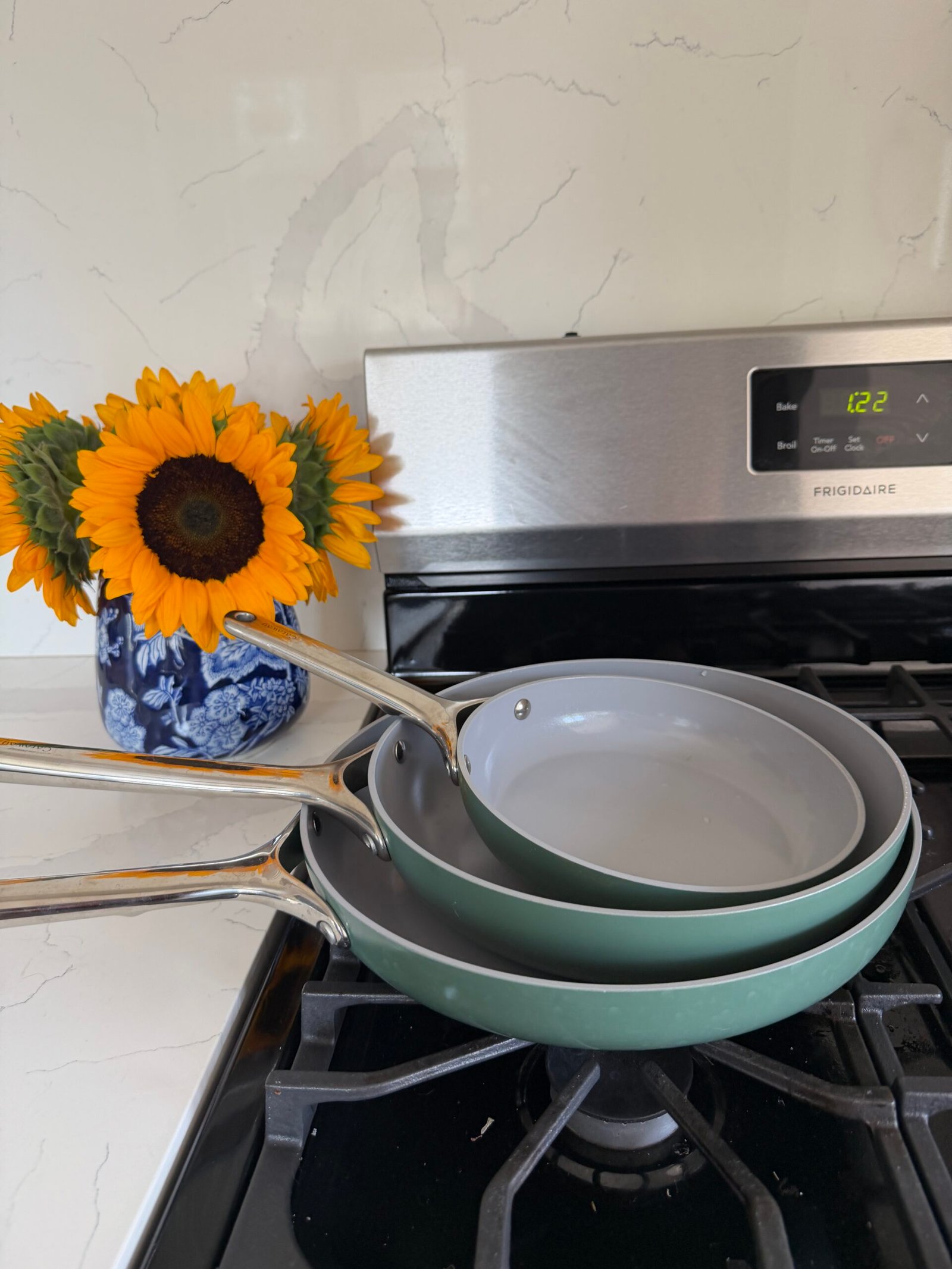 Three nonstick frying pans stacked on a stovetop next to a blue vase with sunflowers. The stove clock displays 12:21.