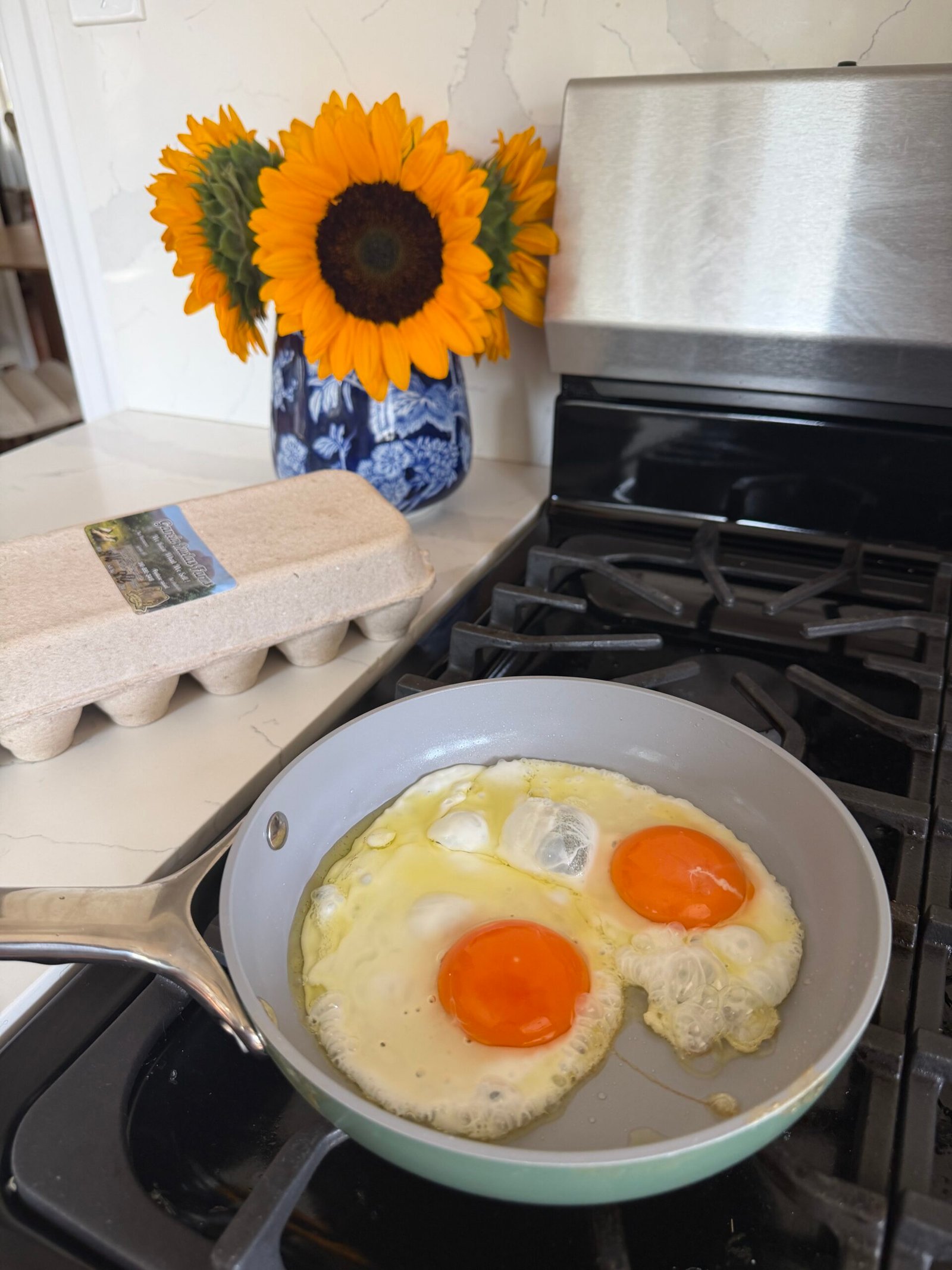 Two eggs are frying in a skillet on a stove, with a carton of eggs and a blue vase holding sunflowers in the background.