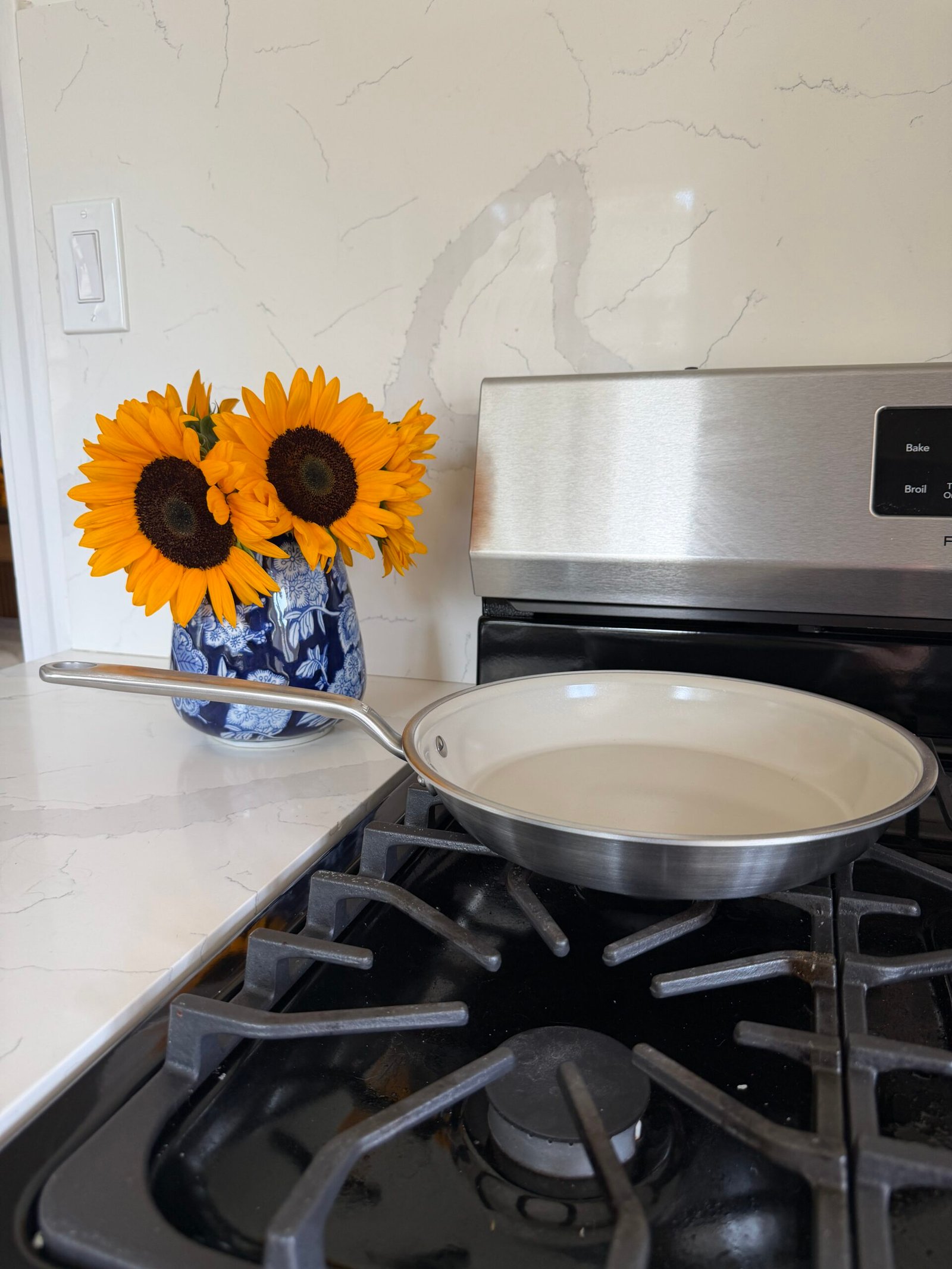 A stainless steel skillet sits on a gas stove next to a blue vase of sunflowers on a white marble countertop.