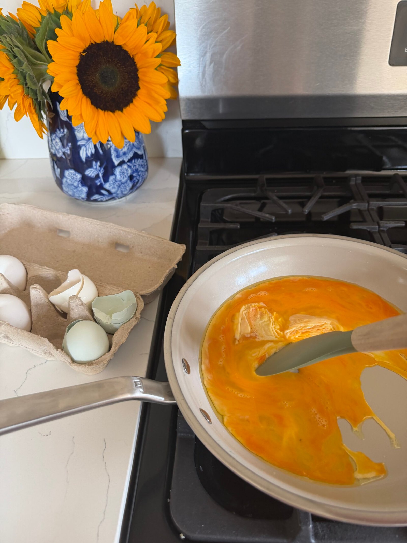 A person is scrambling eggs in a pan on a stove next to an open egg carton with shells and a vase of sunflowers.