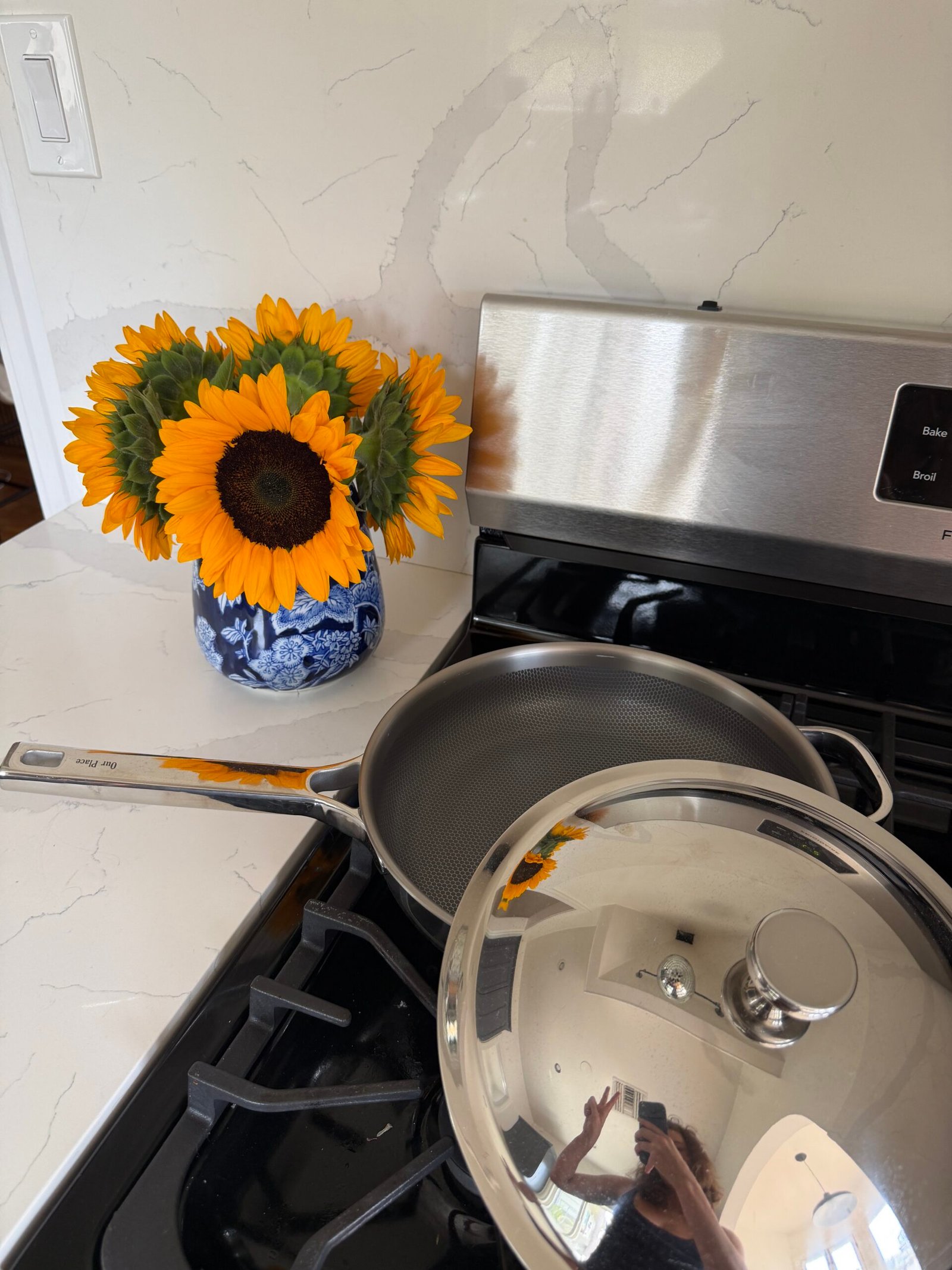 A vase of sunflowers on a kitchen counter next to a stove with a pan and a shiny lid, reflecting a person taking the photo.
