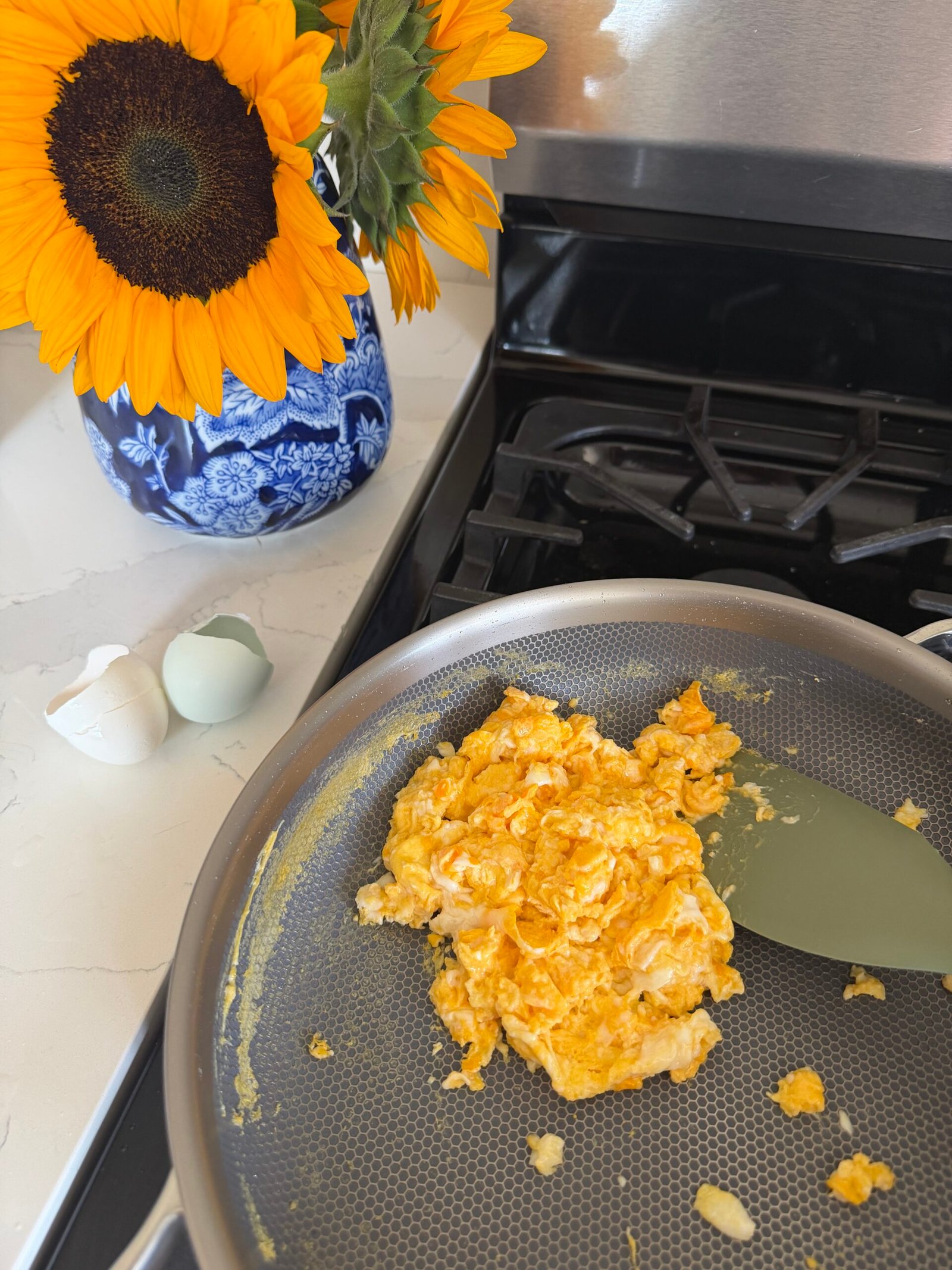 Scrambled eggs cooking in a non-stick pan on a stove, with a spatula, broken eggshells, and a vase of sunflowers on a countertop nearby.