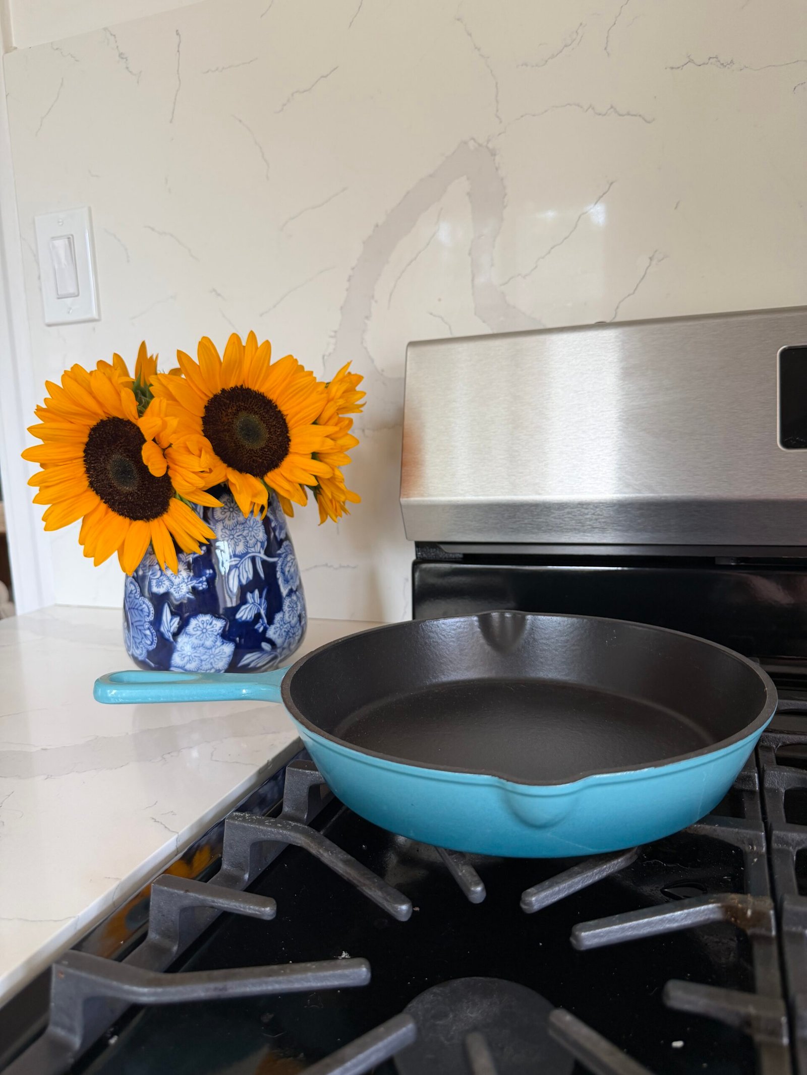 A blue cast iron skillet sits on a stove next to a vase of sunflowers on a white countertop with a marble backsplash.