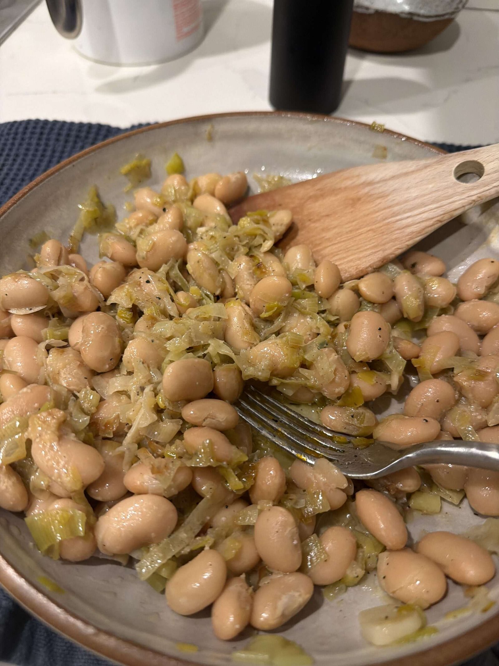 Plate of cooked white beans mixed with sautéed leeks, seasoned with black pepper. A wooden spatula and metal fork are on the plate.