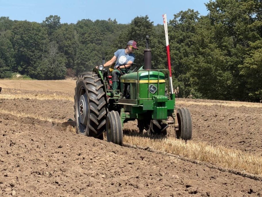 A woman in a ball cap and sunglasses drives a green John Deere tractor pulling a plow, turning a furrow beside standing stubble. Photo: John Greig