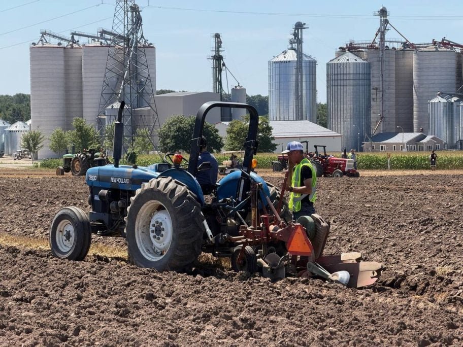 A man in a safety vest stands beside a blue New Holland tractor and plow in a plowed field, with tall grain bins and elevator legs behind. Photo: John Greig